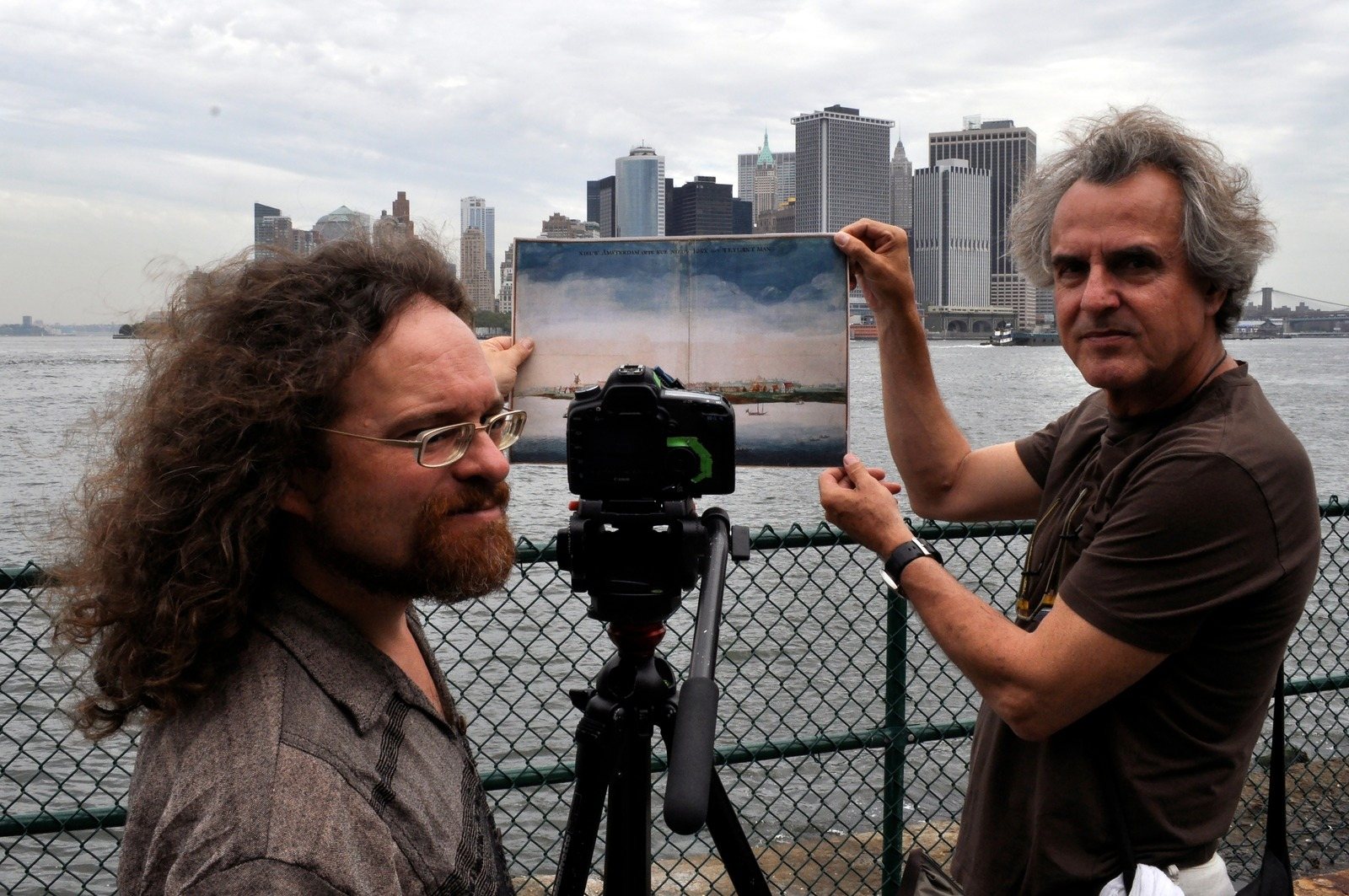 Two men hold up a framed photograph of the Manhattan skyline beside a camera on a tripod, the real skyline across the water behind them