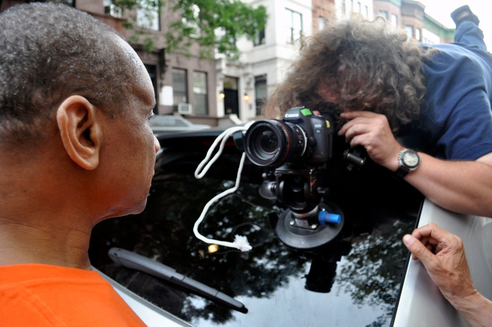 A cameraman leans over a car roof to film Tony Ndogo, seen from behind in an orange shirt, on a New York street