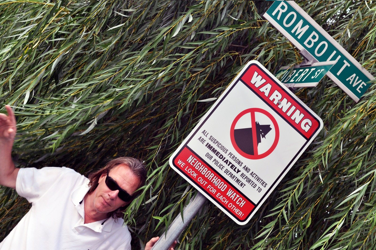 A man in sunglasses leans out of tall grass beside a Neighborhood Watch warning sign and a street sign for Rombout Ave