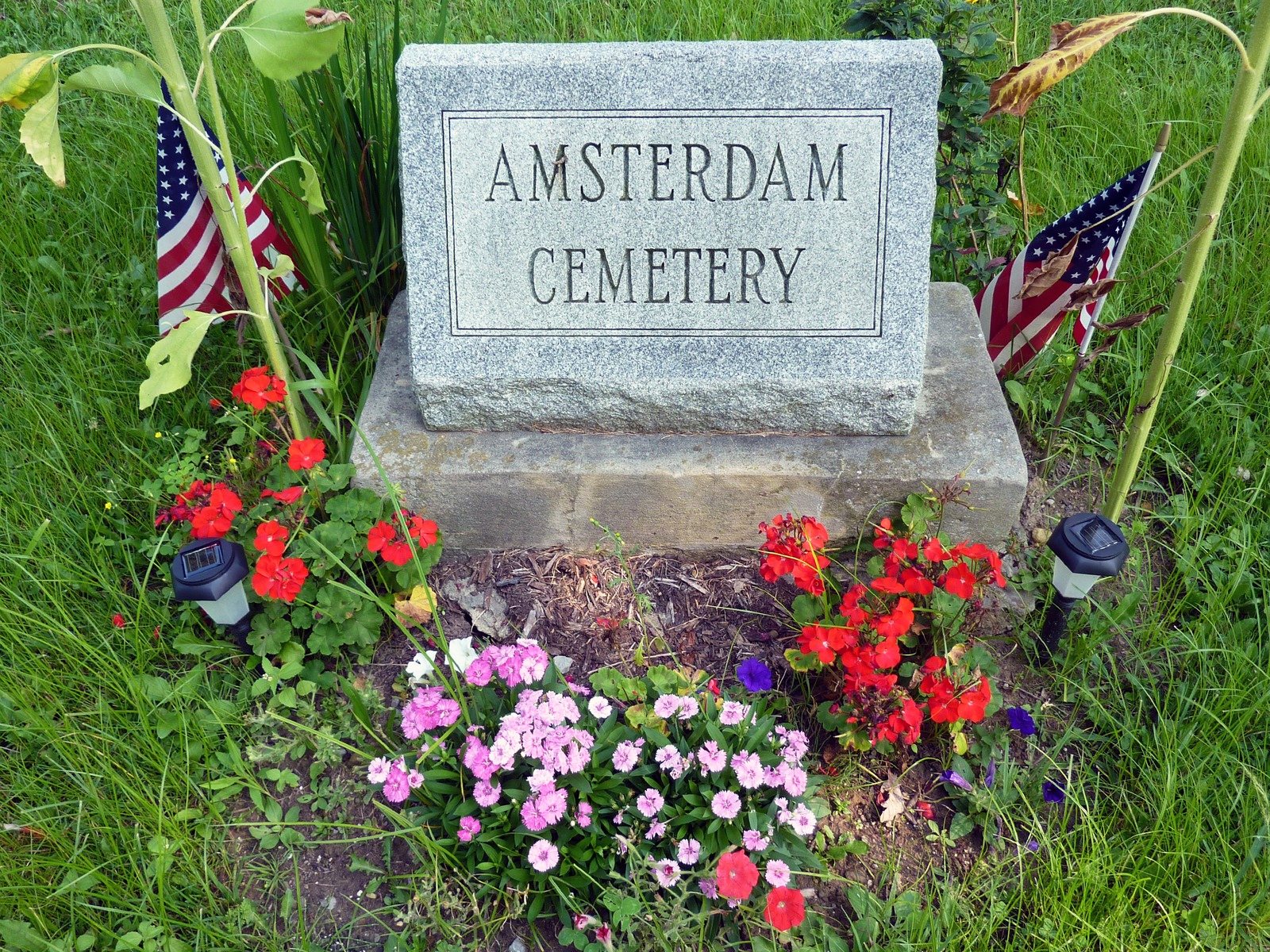 A granite stone reading Amsterdam Cemetery among red and pink flowers and two small American flags