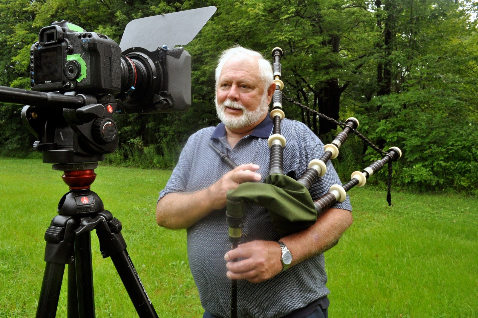 Lee McCoy, white-bearded, holds a set of bagpipes on a lawn beside a camera on a tripod