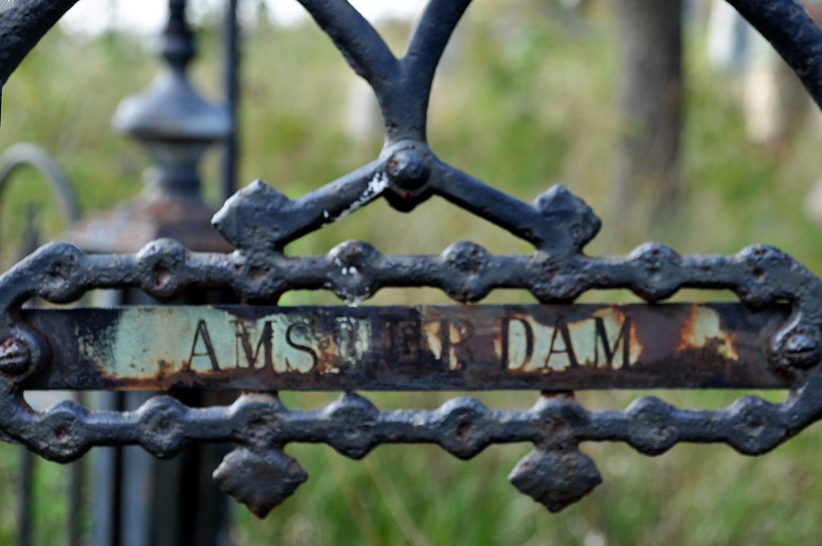 A rusted iron gate sign reading Amsterdam, green leaves behind