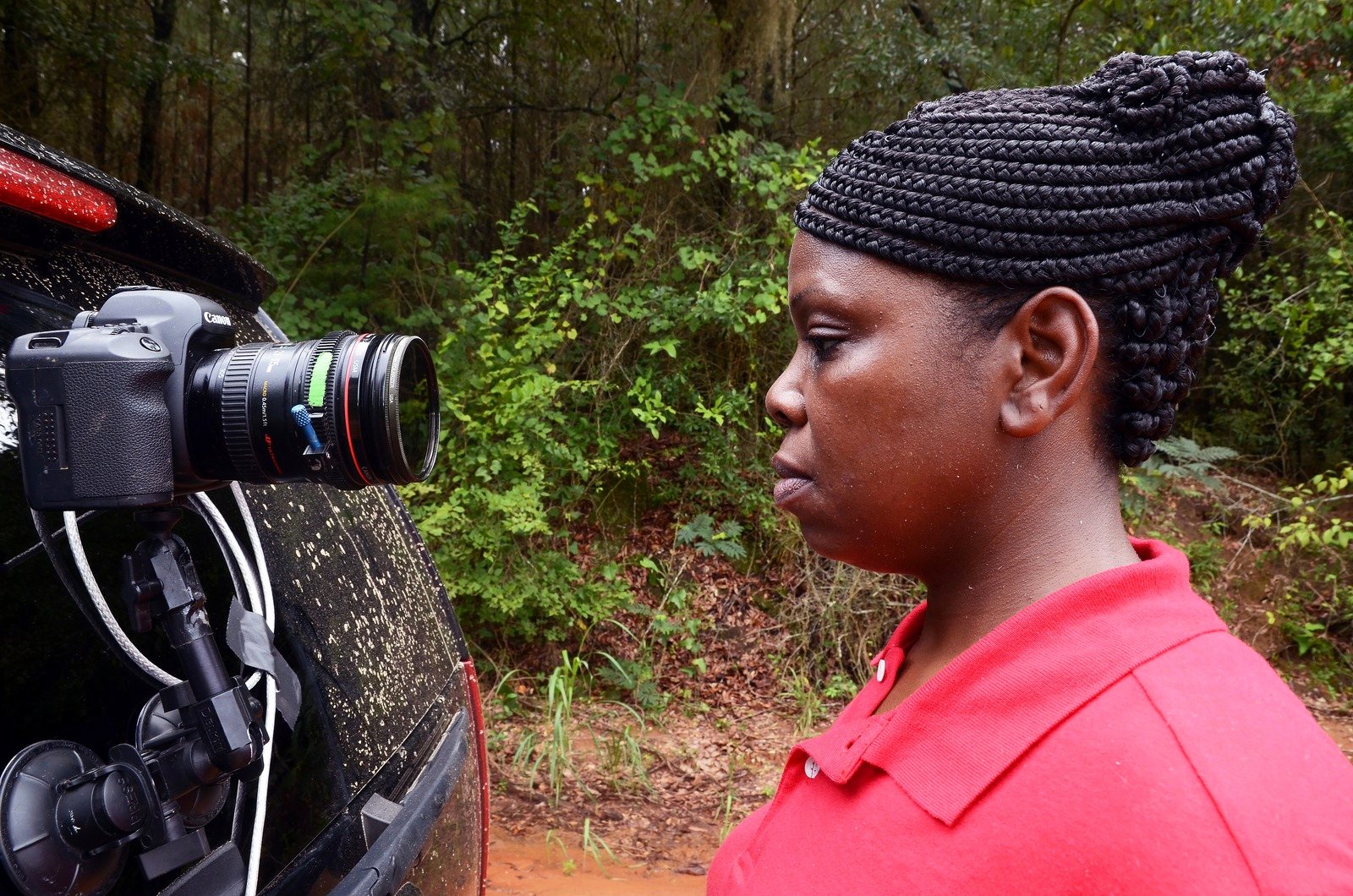 A woman in a red shirt and braided hair stands by a car with a camera mounted on its side, woods behind