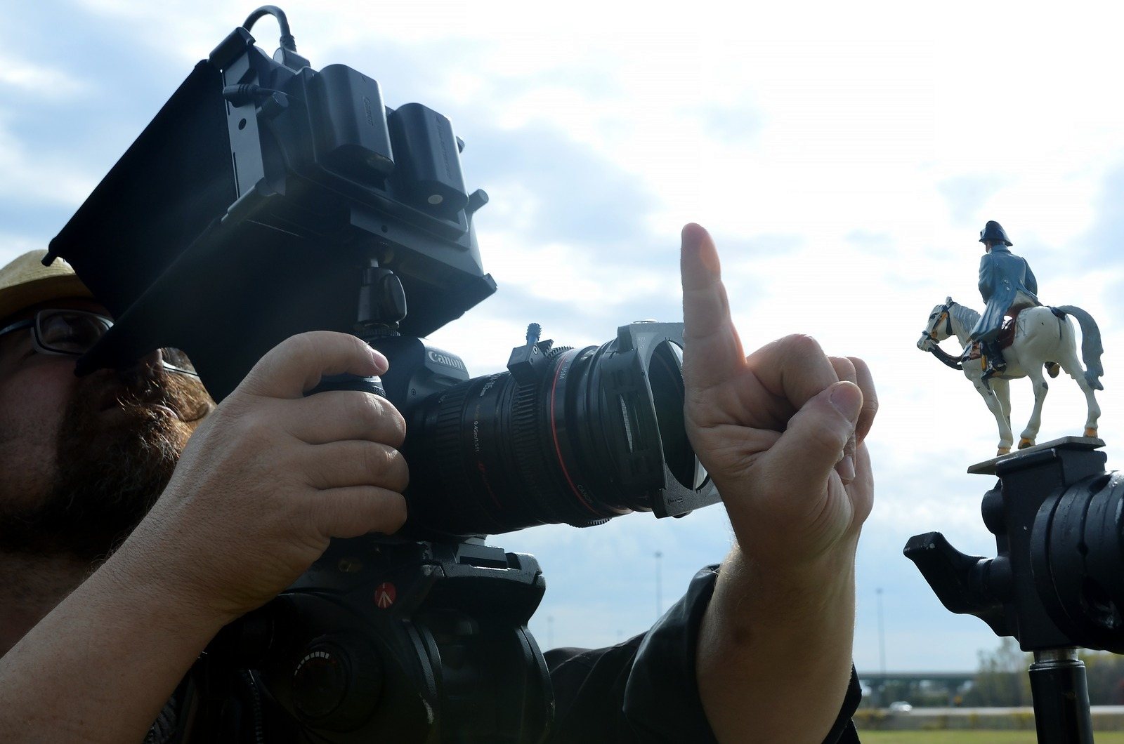 A cameraman with a large camera on his shoulder, a raised finger, and an equestrian statue on a column behind