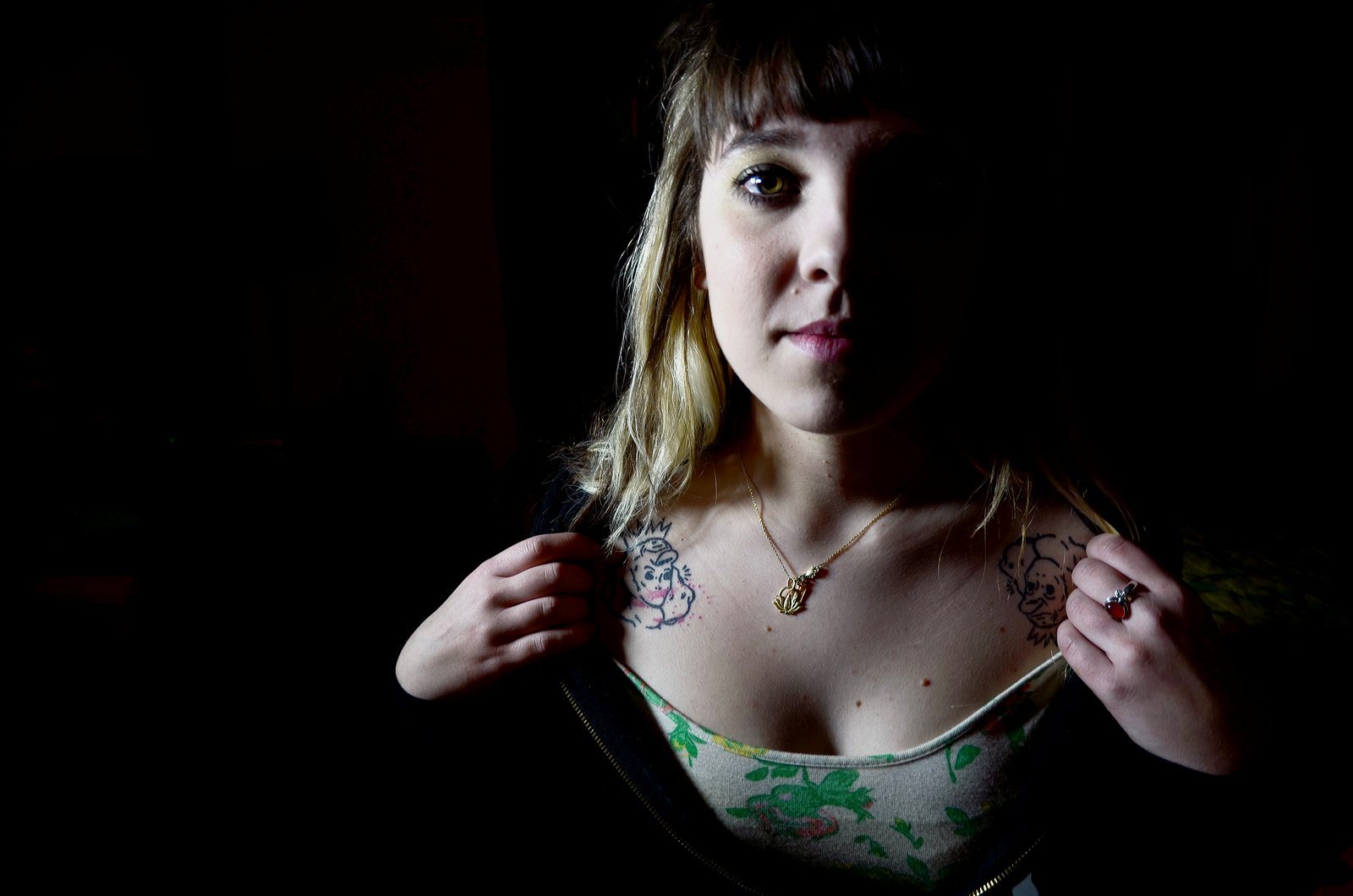 Chesney Sherman, half lit against a dark background, holds open her top to show tattoos below her collarbones