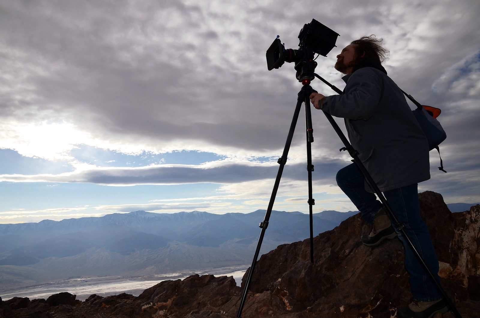 Ben Wolf at a camera on a tripod on a rocky ridge at Dante’s View, above Death Valley under heavy clouds