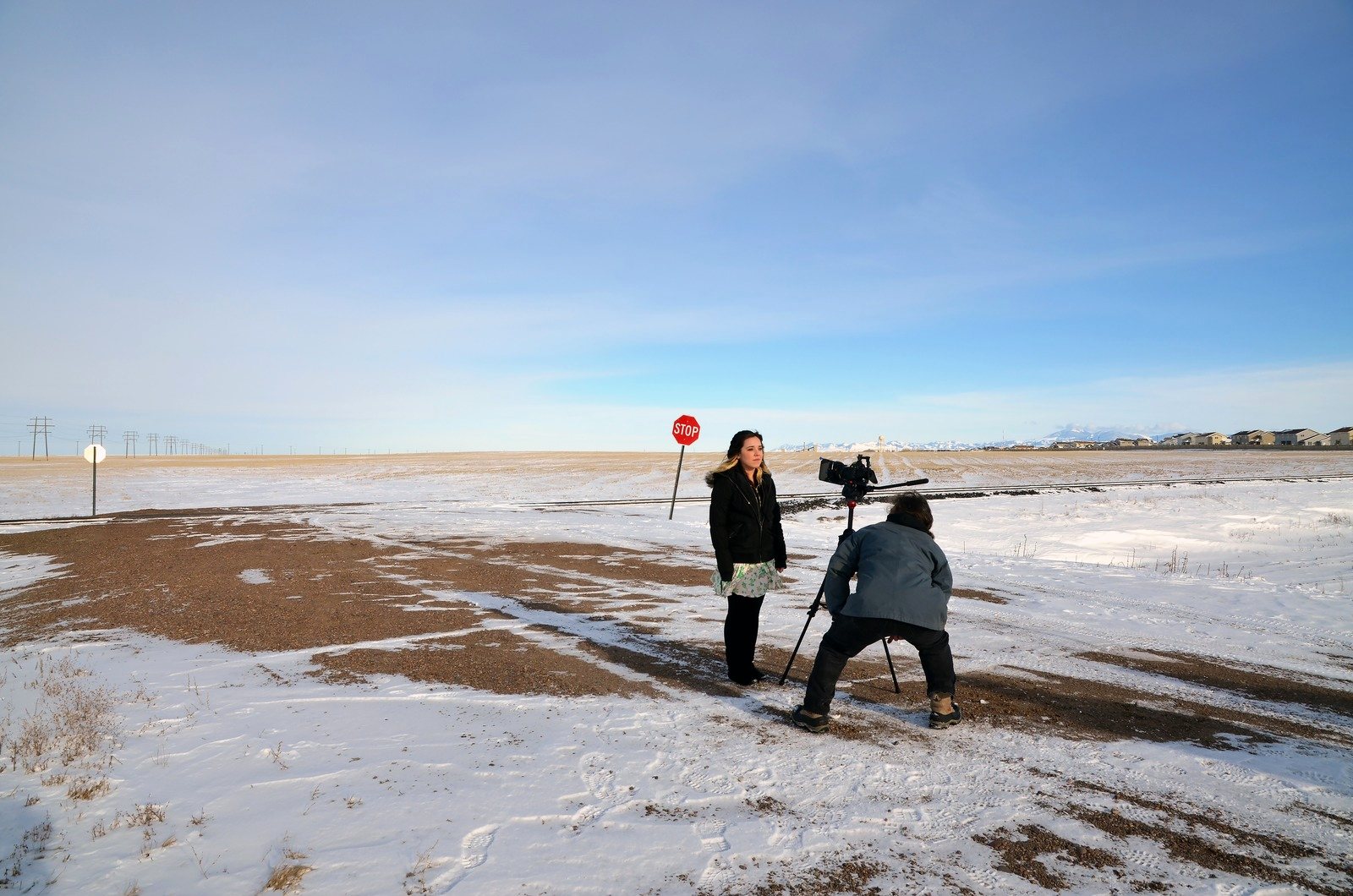 A young woman and a cameraman with a tripod on a snowy plain beside a stop sign, mountains far off