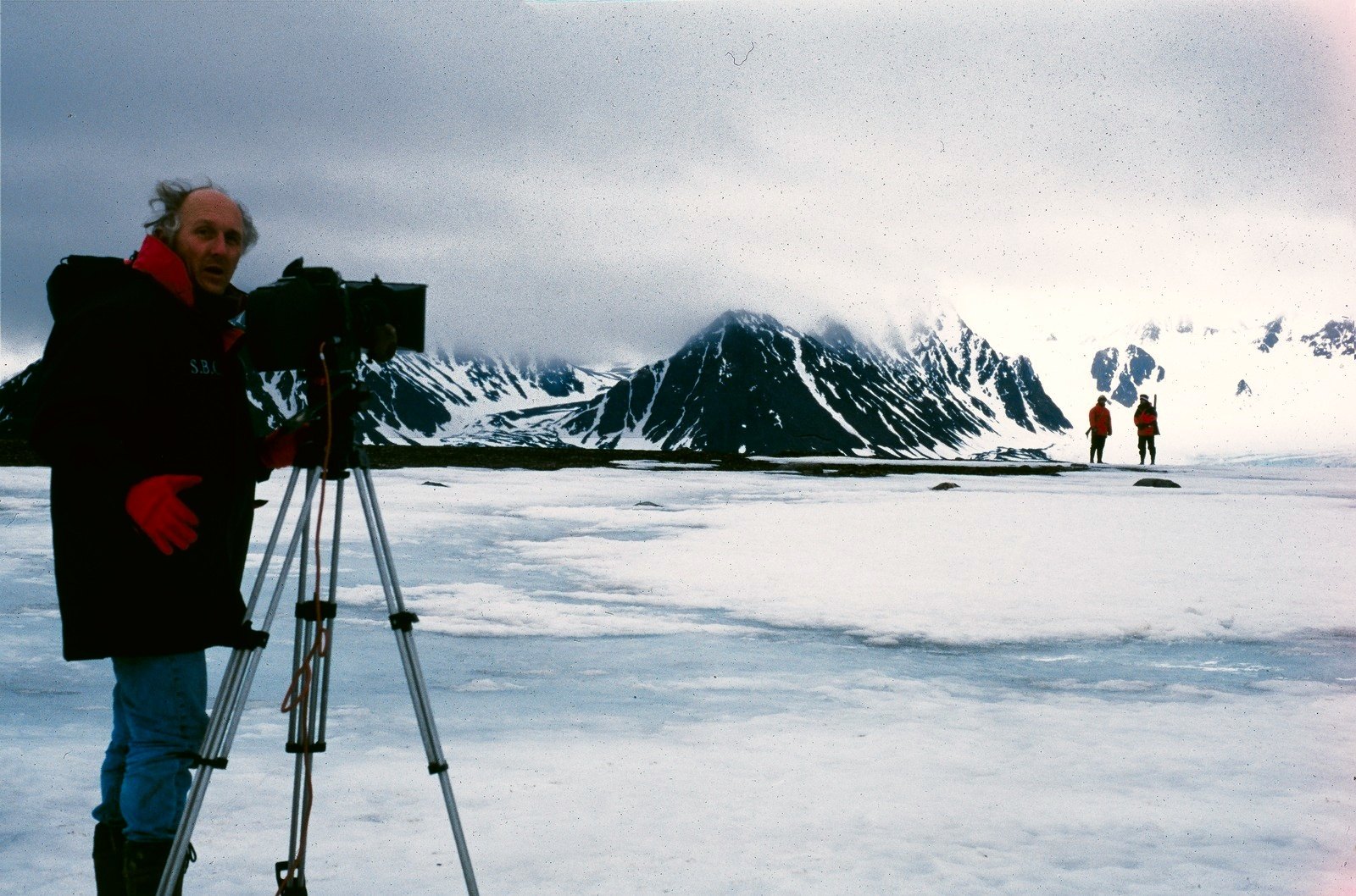 A cameraman in a red and black parka at a camera on a tripod on snow, dark mountains and two figures in the distance