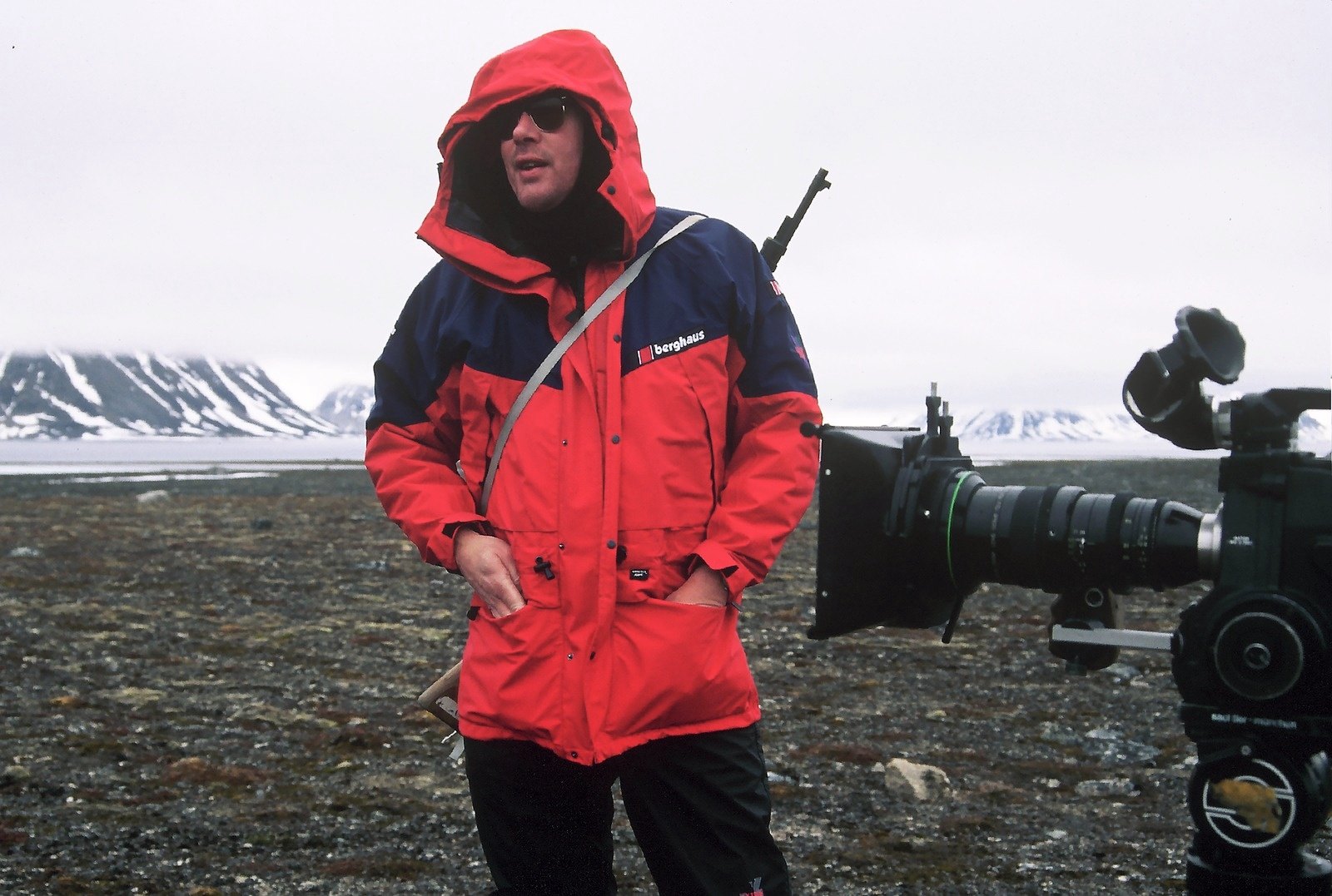 A man in a red hooded parka and sunglasses on stony ground, a film camera beside him