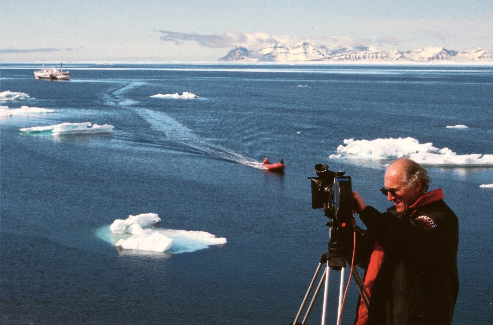 A man at a camera on a tripod on a ship’s deck, blue water with ice floes and a small red boat, snowy mountains far off