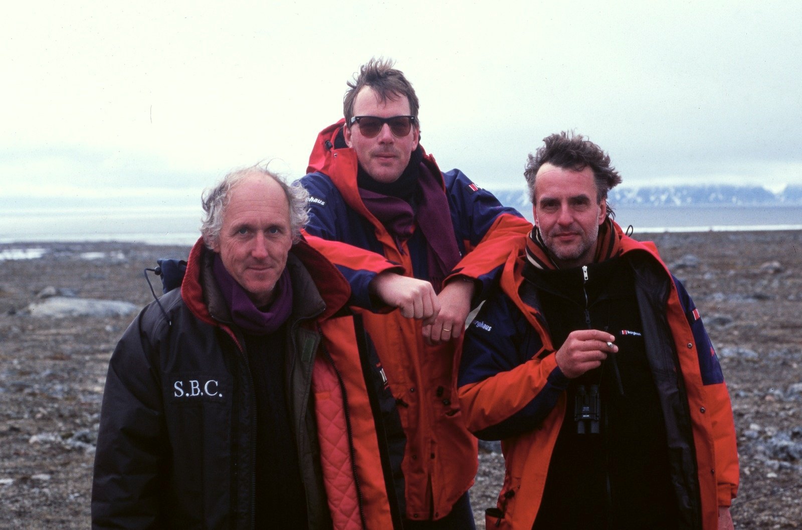 Three men in parkas close together on a stony shore, the sea and a low coast behind