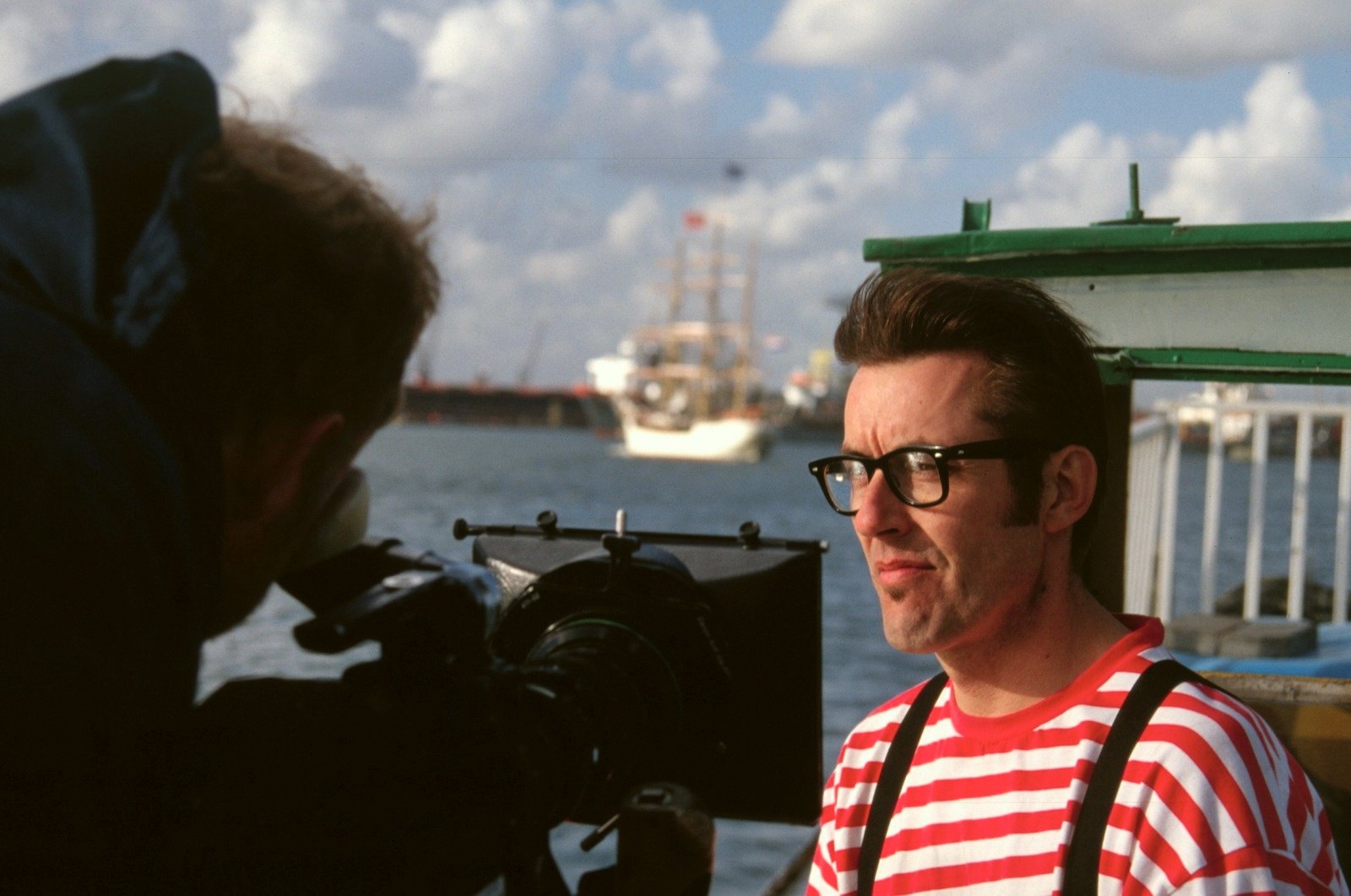 A man in a red and white striped shirt and braces beside a camera, a tall ship in the harbour behind