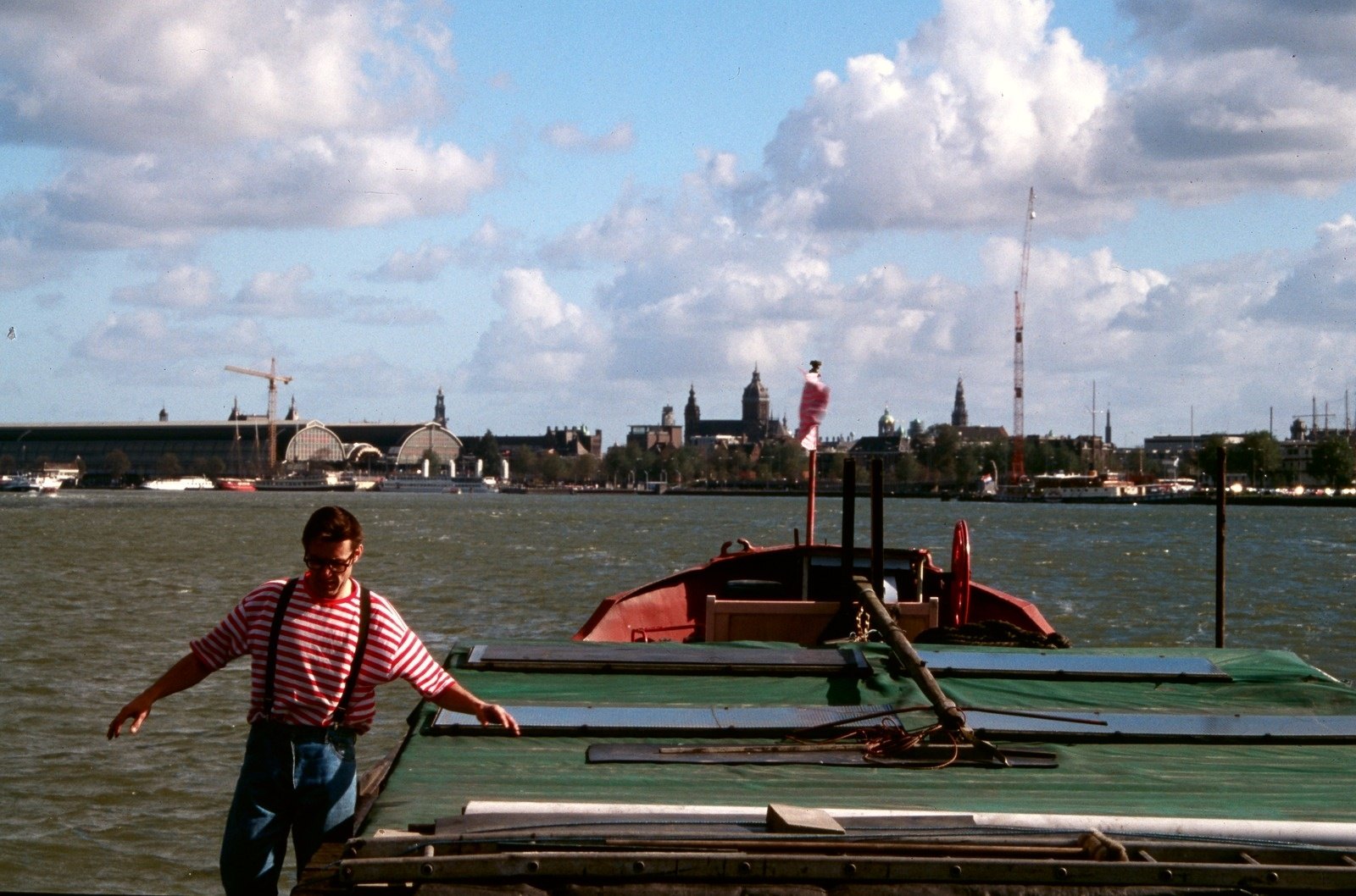 A man in a striped shirt on the deck of a barge, the Amsterdam waterfront under big clouds behind