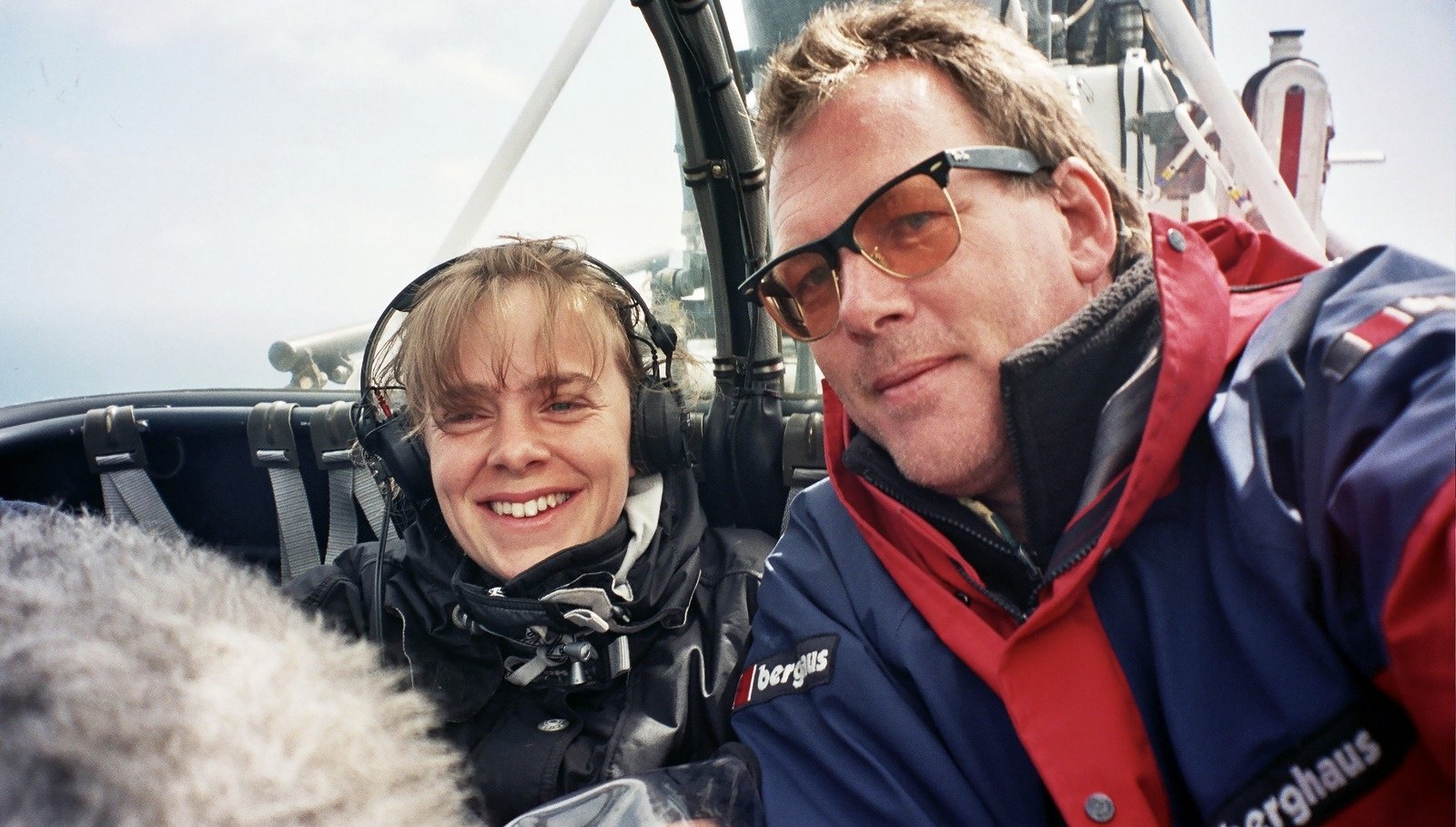A smiling woman with headphones and a man in tinted glasses and a red and blue jacket inside a helicopter