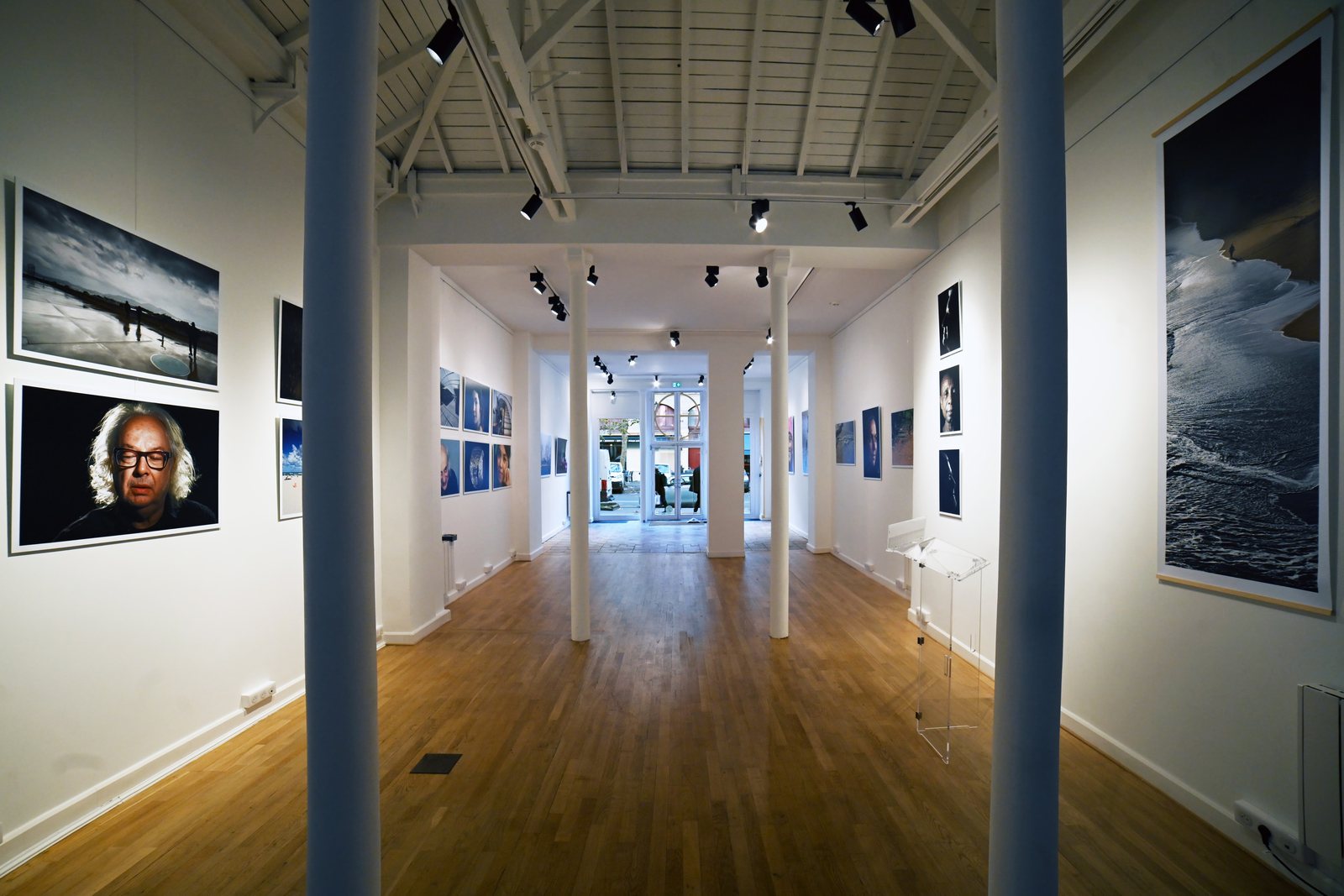 A long white gallery with large colour portraits and a tall seascape on the walls