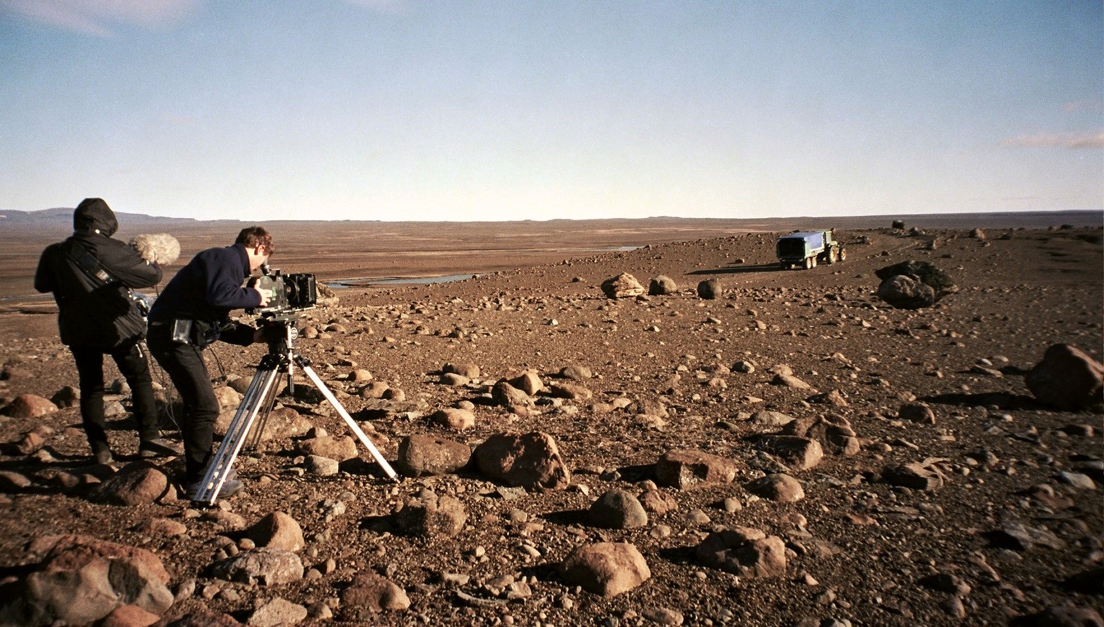 Two crew members with a camera on a tripod on a wide plain of brown stones, a vehicle far off