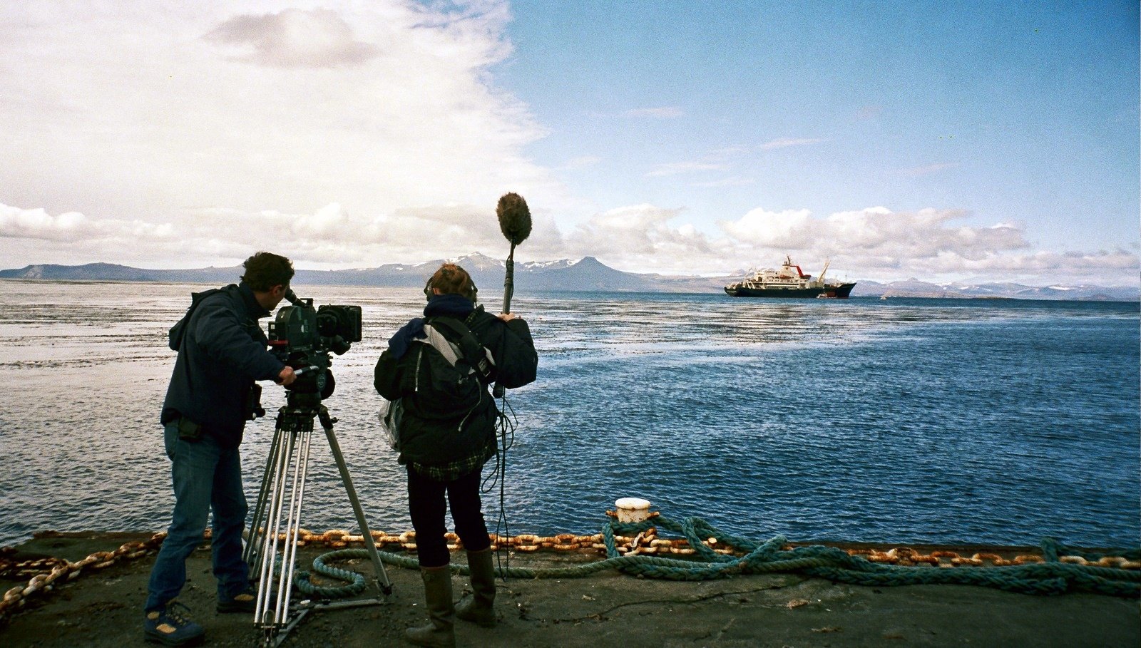 A cameraman at a tripod and a sound recordist with a boom microphone on a quay, a ship at anchor across the blue water