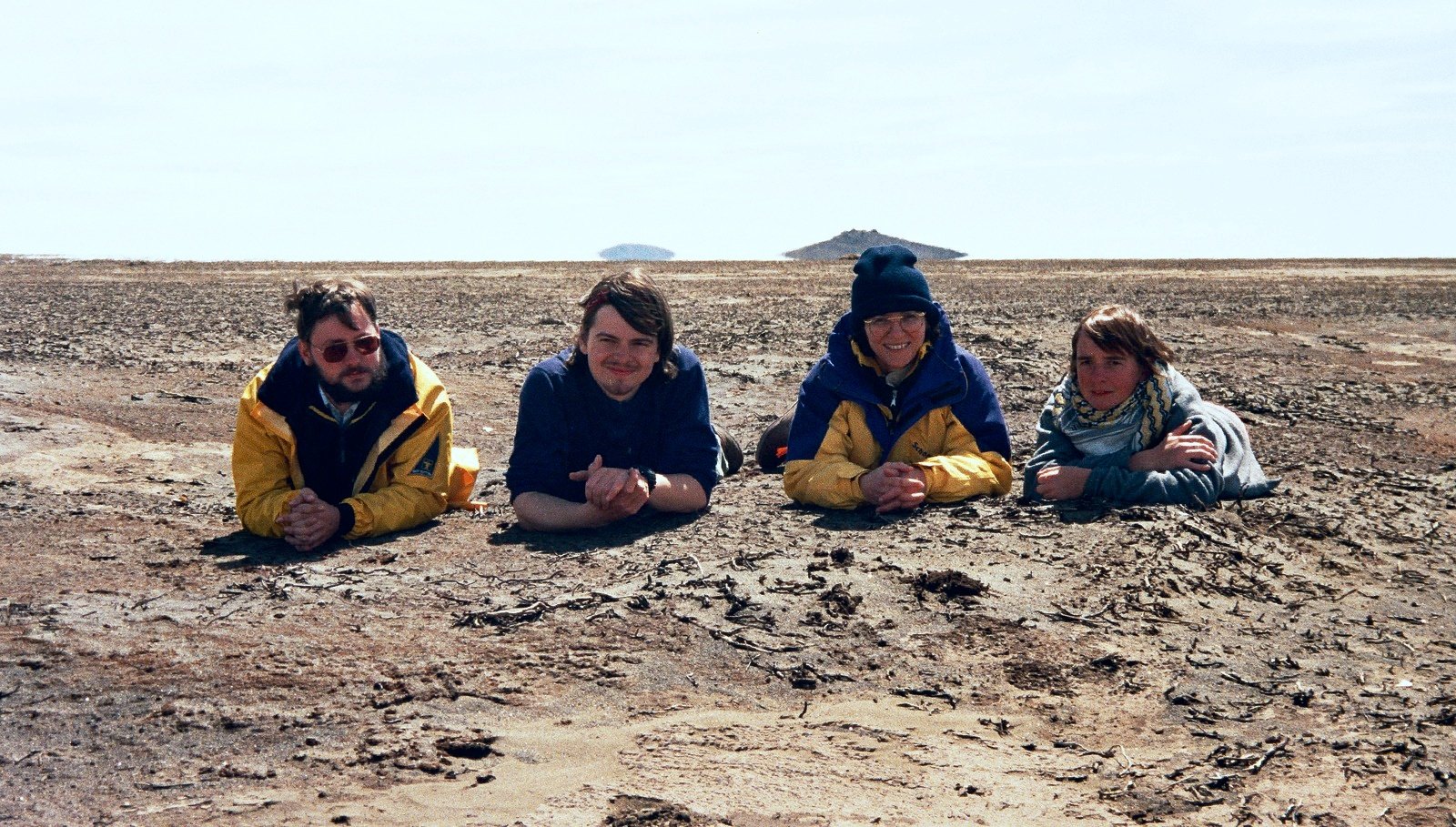 Four people in bright jackets lying side by side on the bare ground, chins on their hands