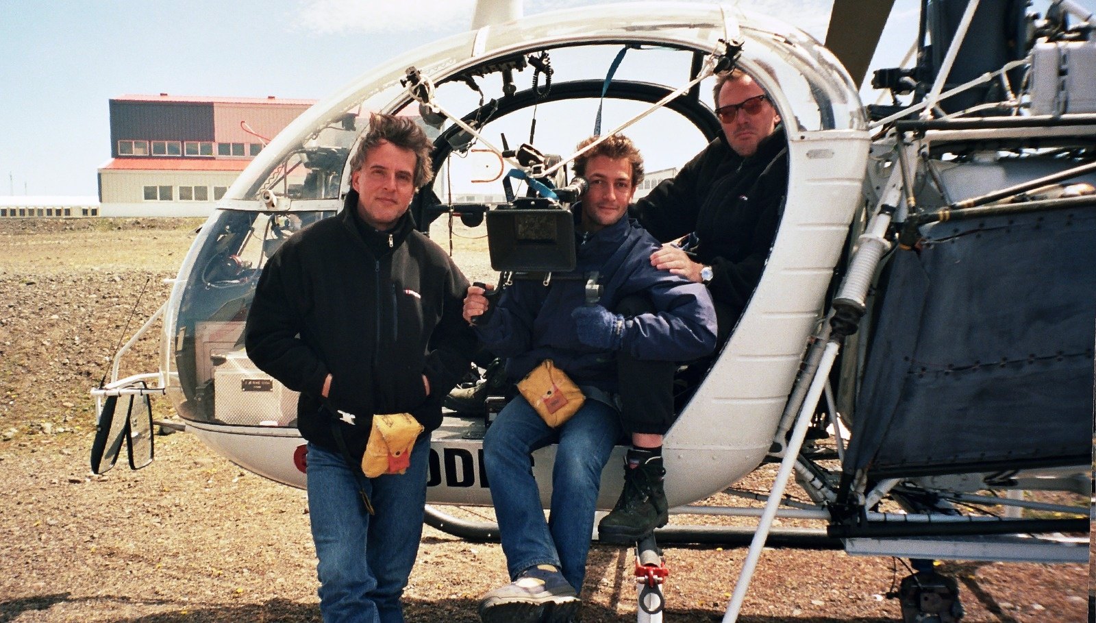 Three men in the open cabin of a small white helicopter, a camera between them