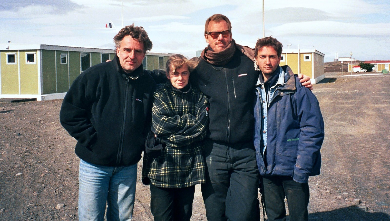 Four people standing close together in front of low prefabricated buildings