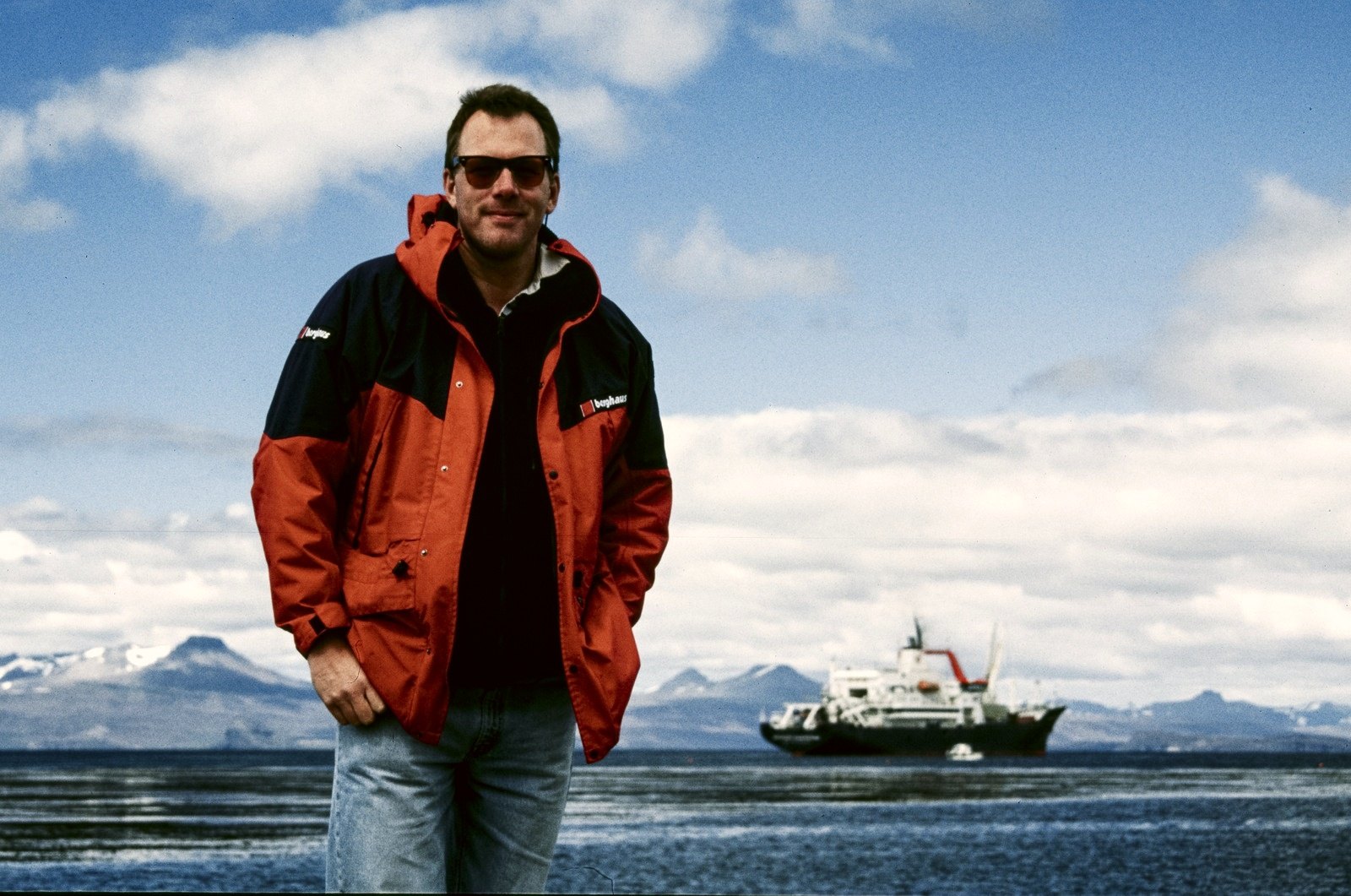 A man in sunglasses and a red and black jacket on the shore, a ship at anchor and snowy mountains behind