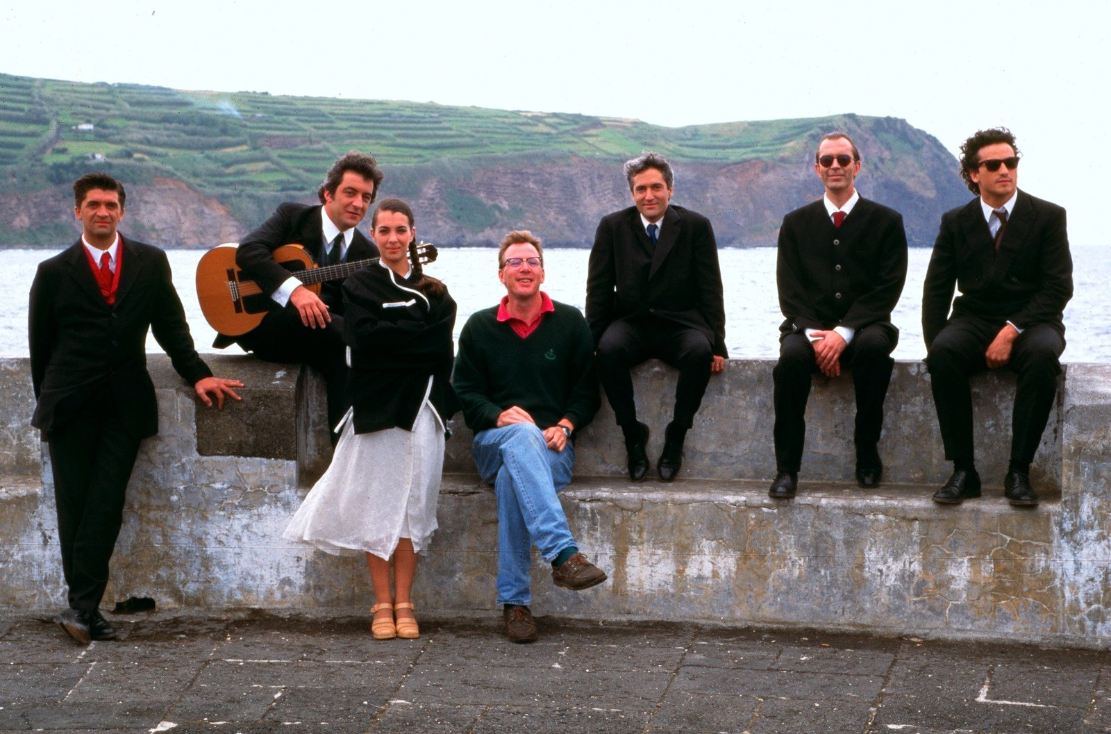 Six men and a woman in a white skirt on a concrete sea wall, green terraced hills across the water