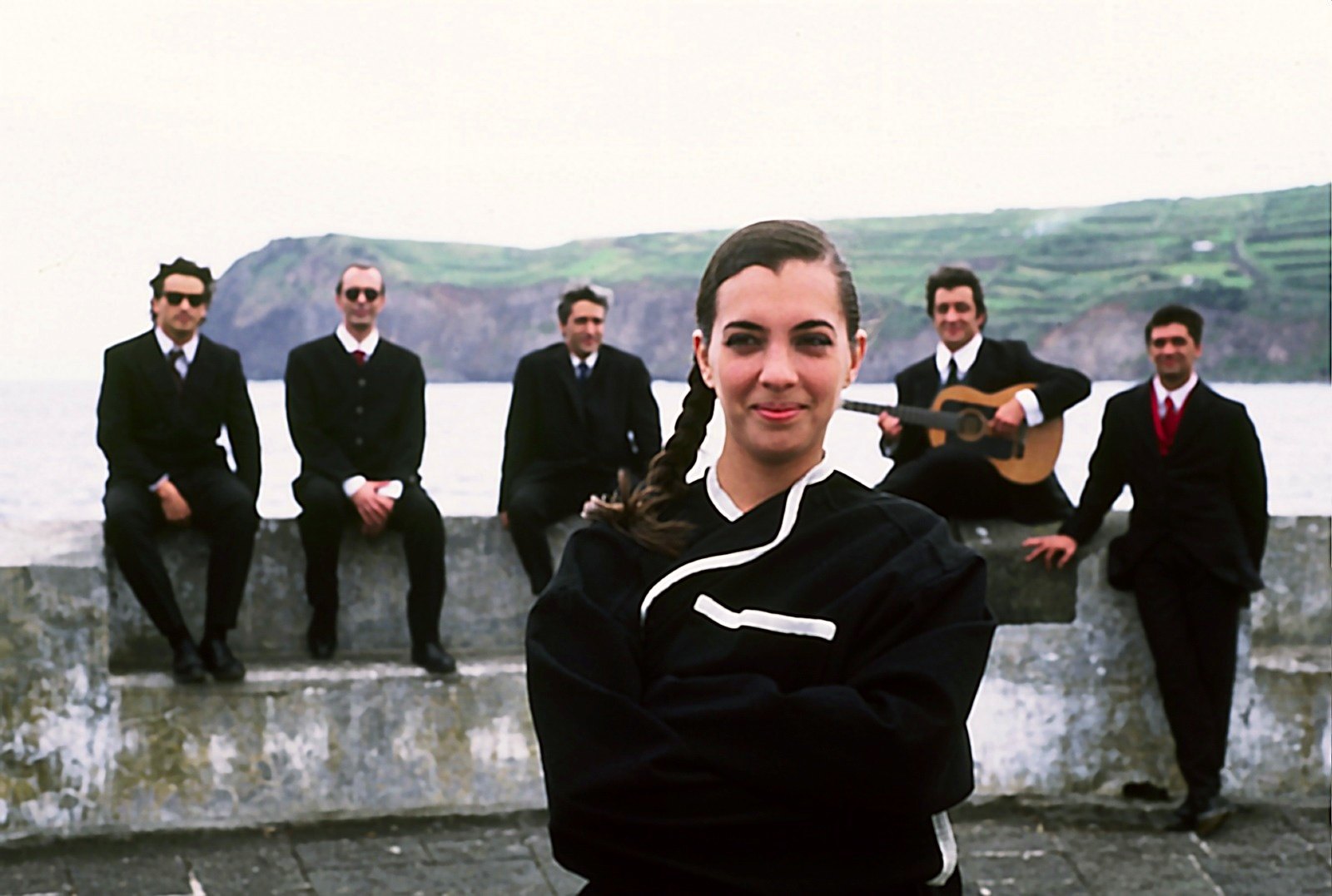 A young woman with a braid in a black jacket in front, five men in dark suits on the sea wall behind her