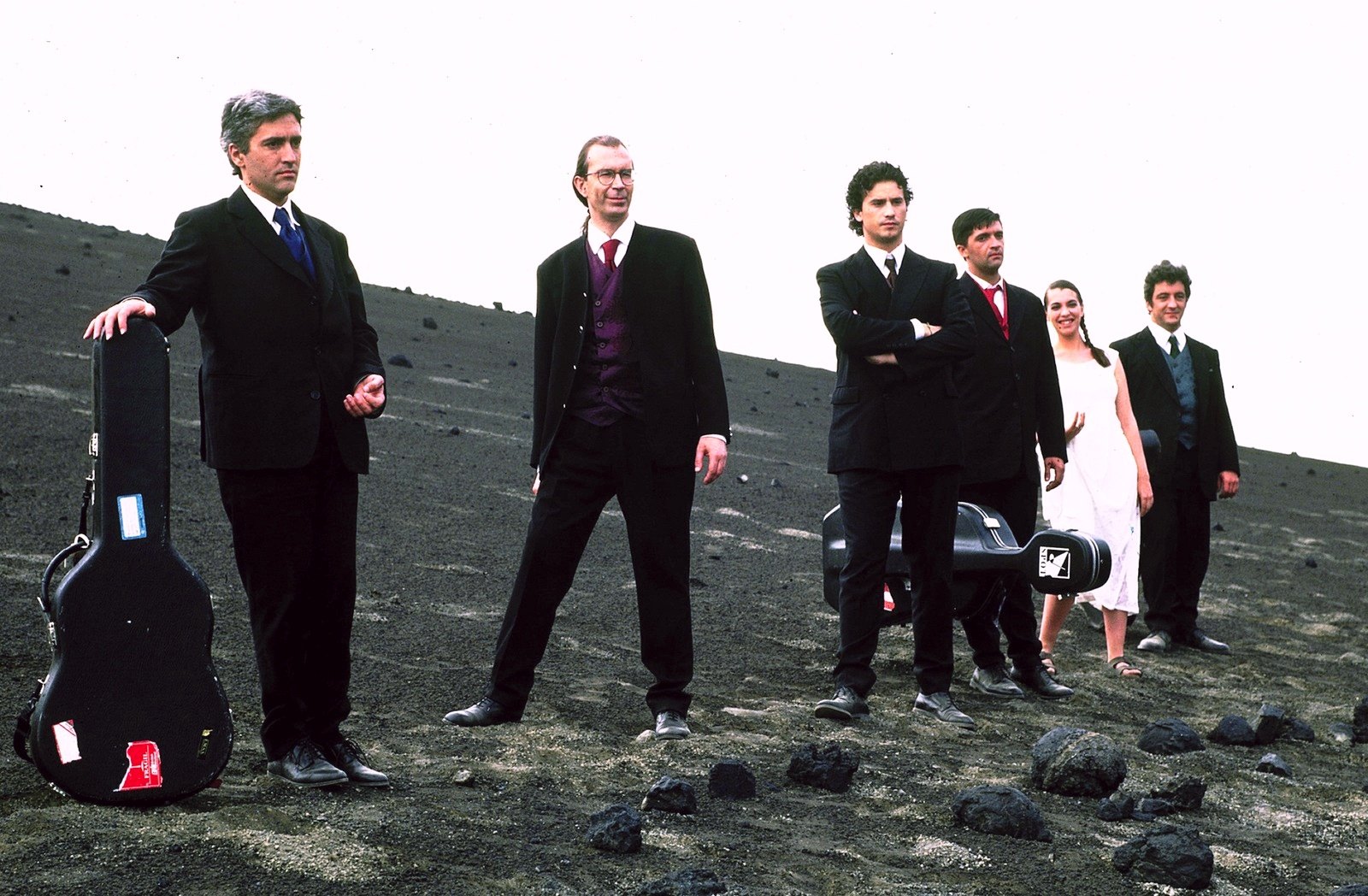 Musicians in dark suits and a woman in white standing on a slope of black volcanic ash, a guitar case in front