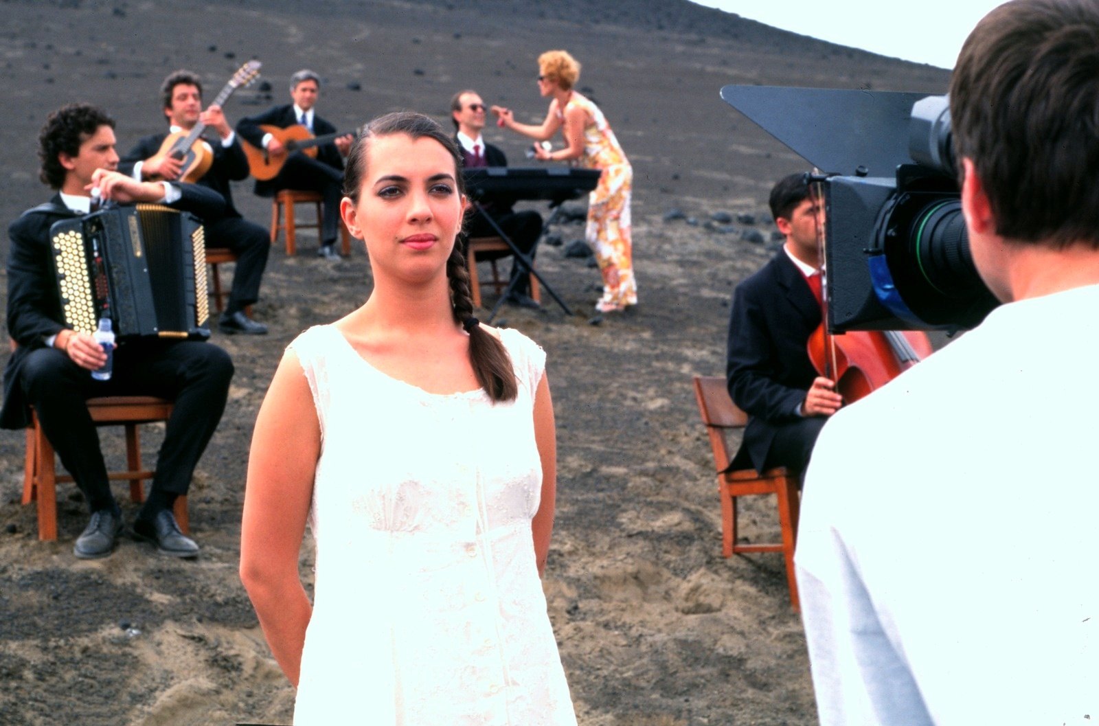 A woman in a white dress faces the camera, musicians seated behind her on the ash; a cameraman in the foreground