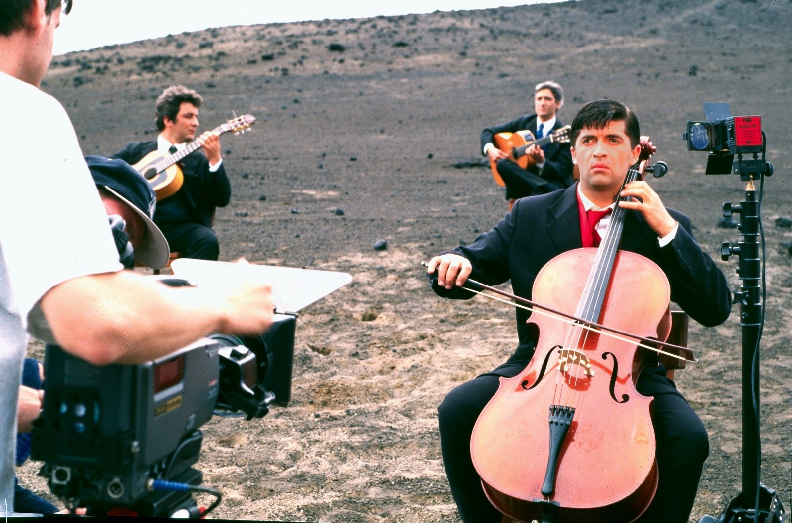 A cellist in a dark suit plays on the ash slope, a video camera in the foreground, guitarists behind