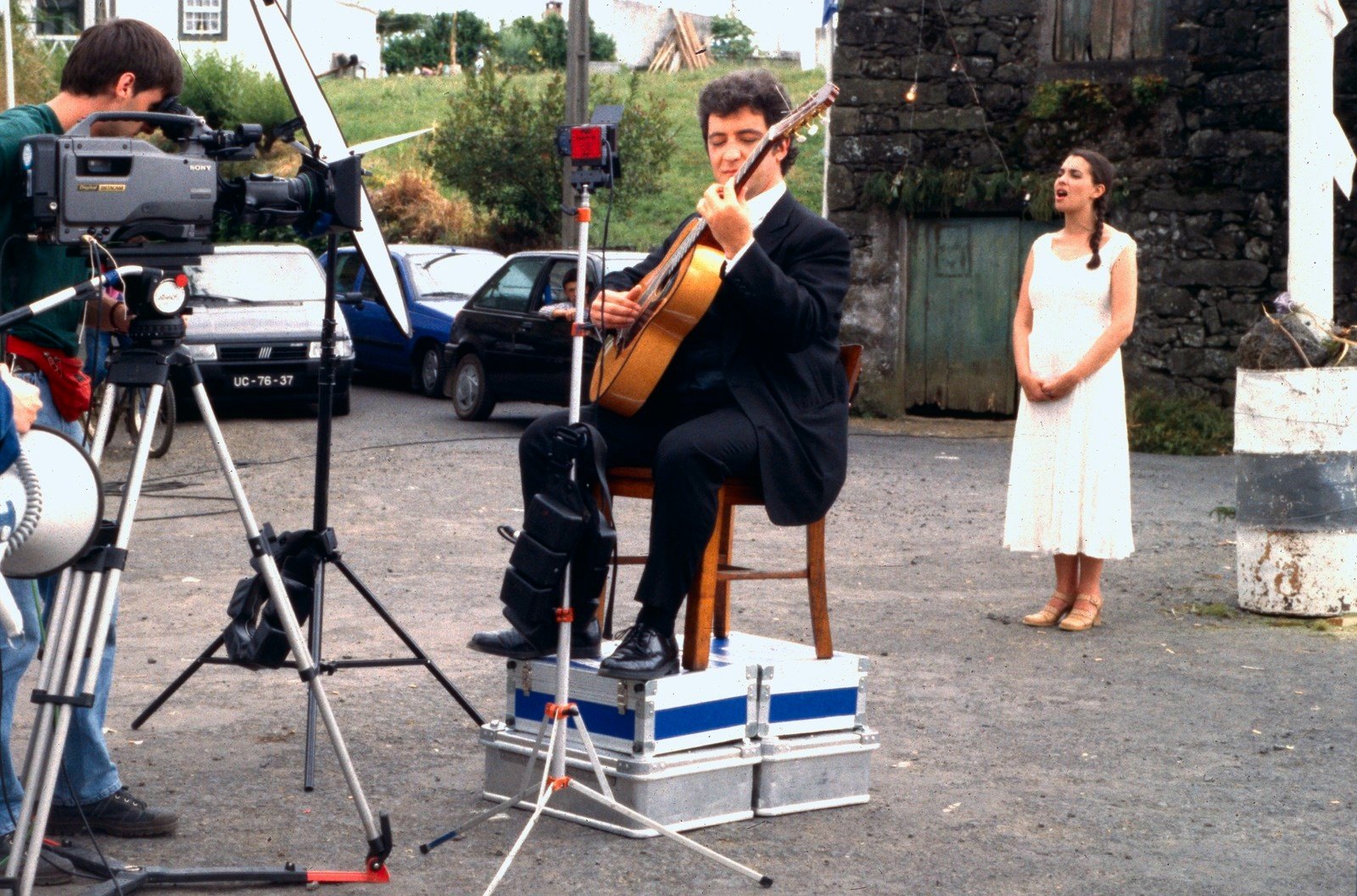 A guitarist seated on equipment cases, a woman in white standing nearby, a camera on a tripod at the left