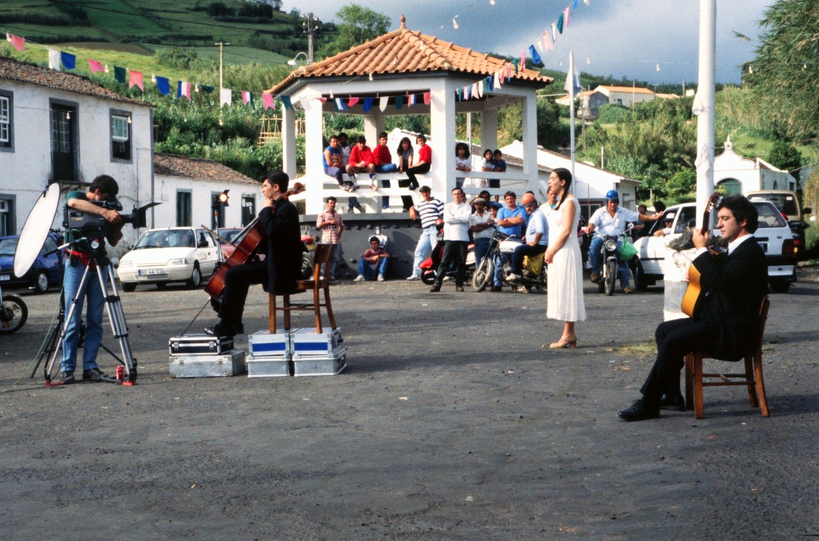 A village square with a bandstand under a tiled roof, musicians and a woman in white performing for the camera, onlookers behind