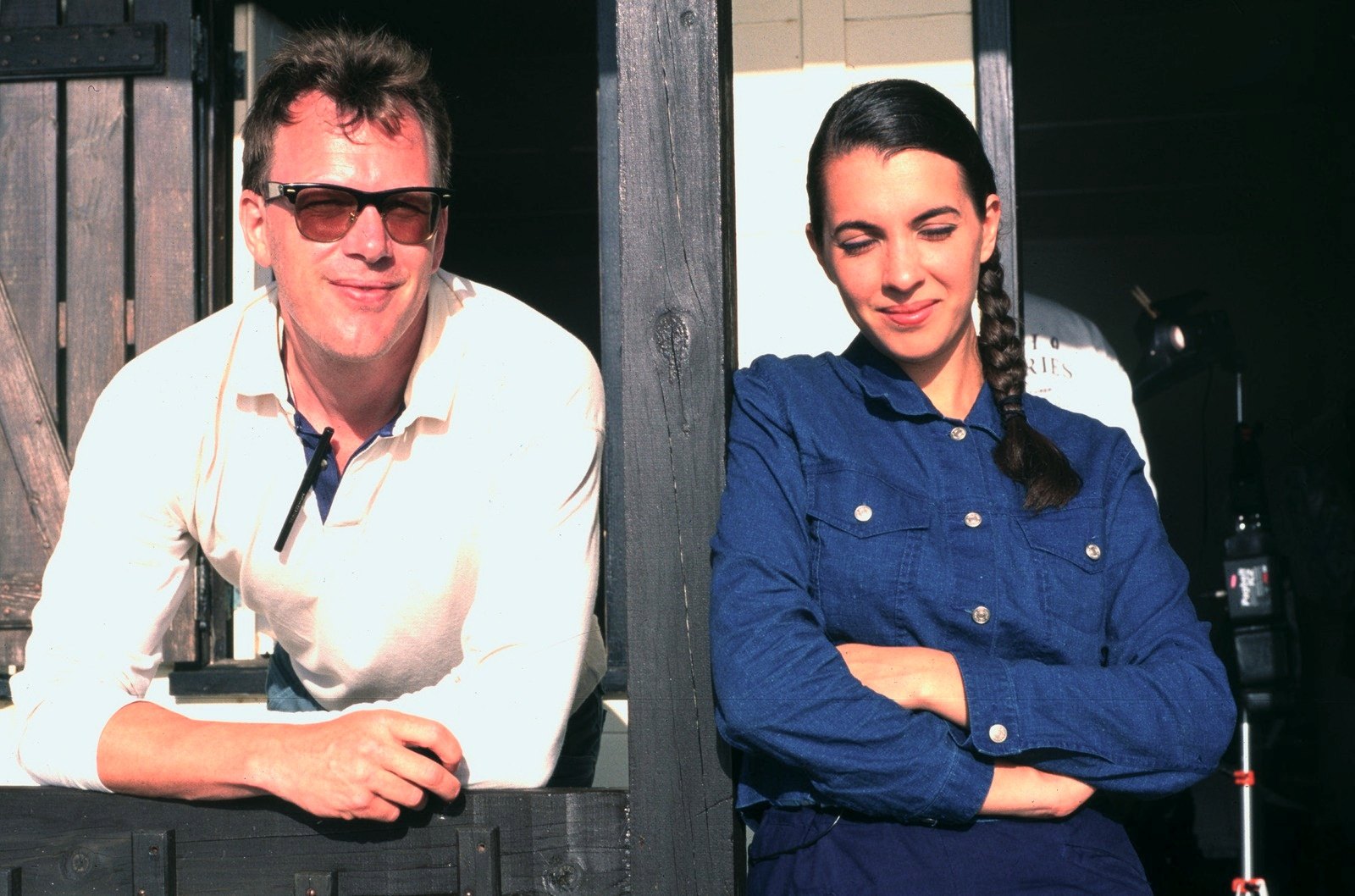 A man in sunglasses and a white shirt and a young woman with a braid in a blue shirt, leaning in a wooden doorway