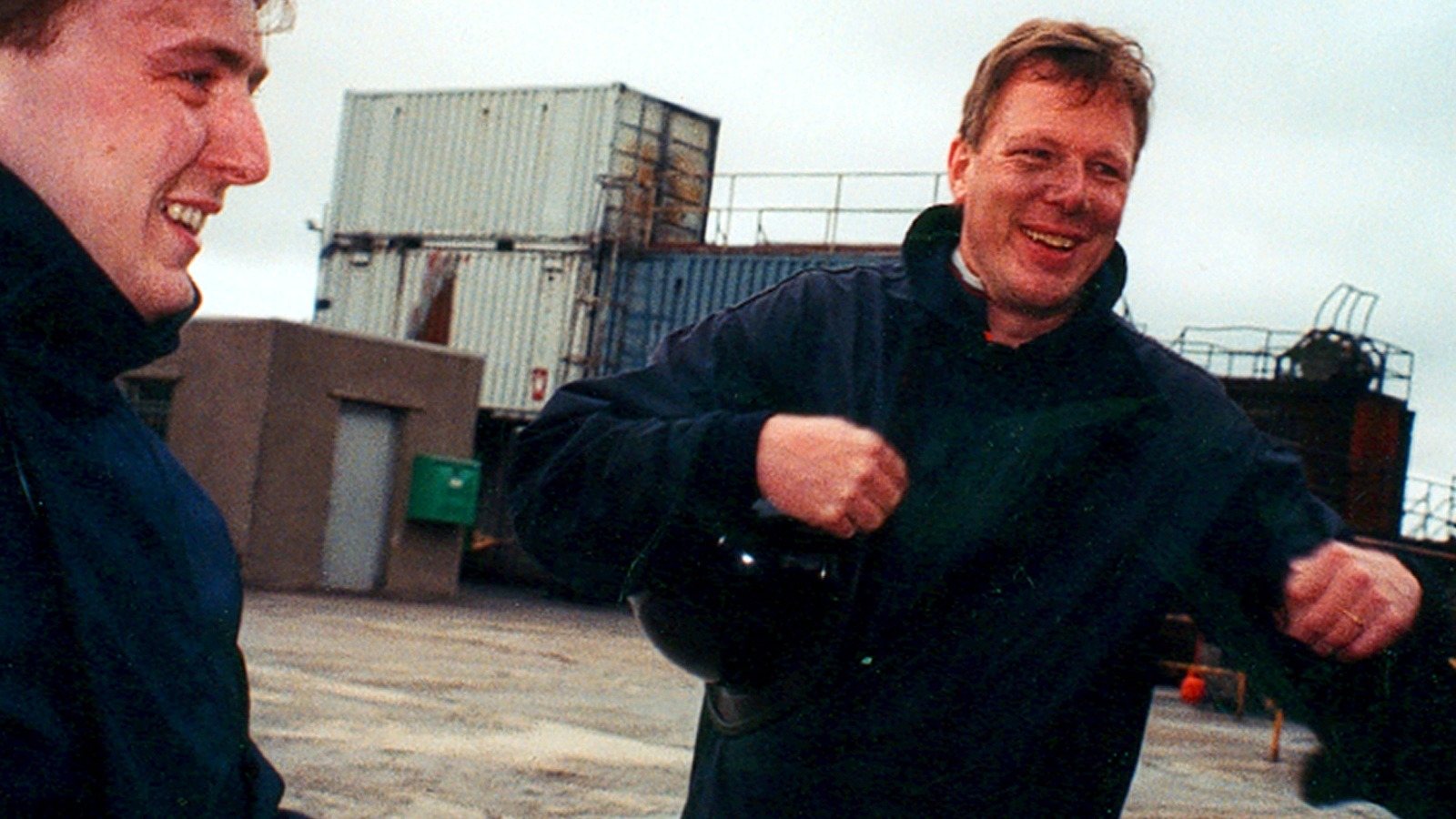 Two men laughing in dark jackets on an industrial yard with containers