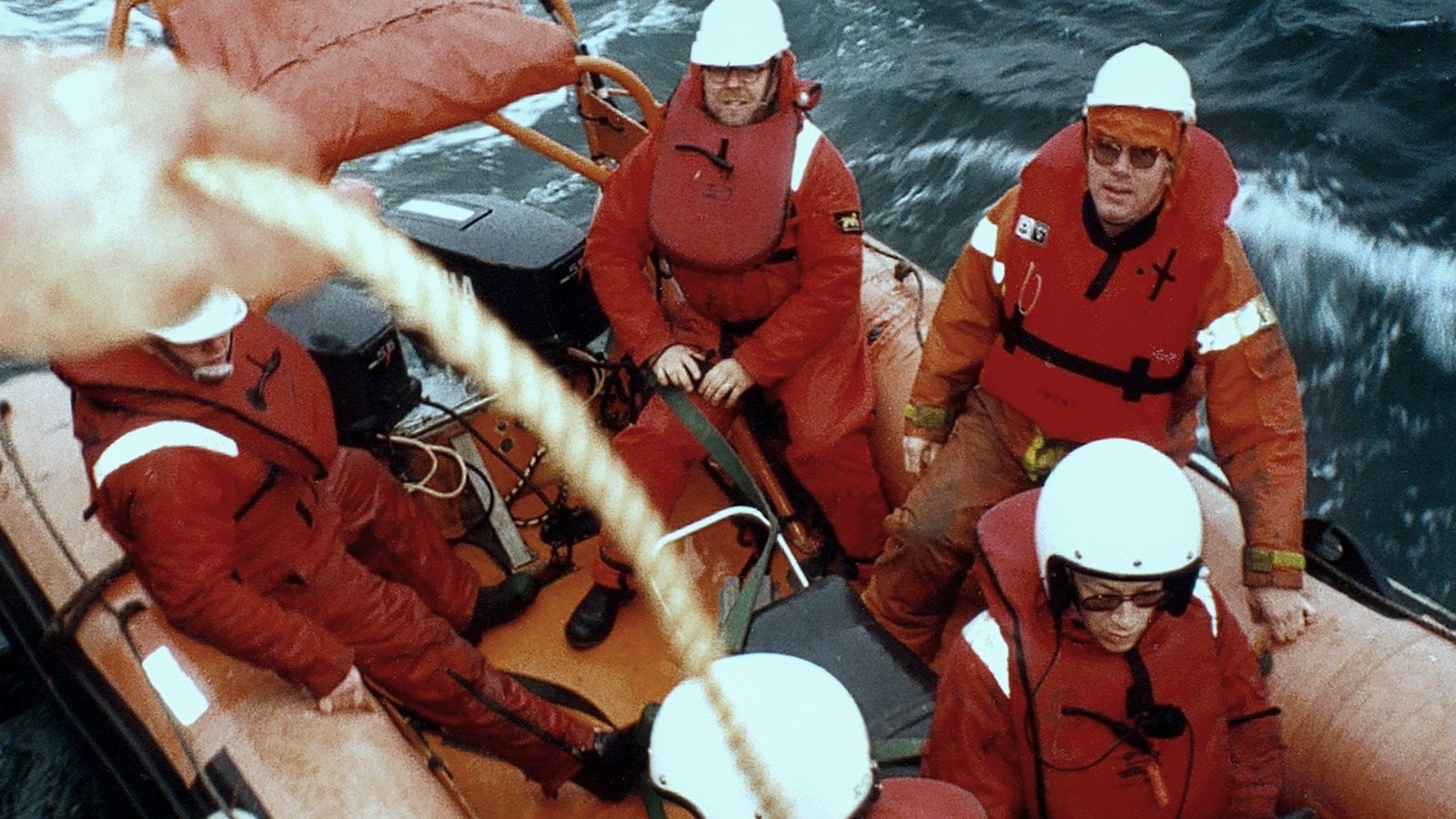 People in orange survival suits and white helmets in an inflatable boat on rough water