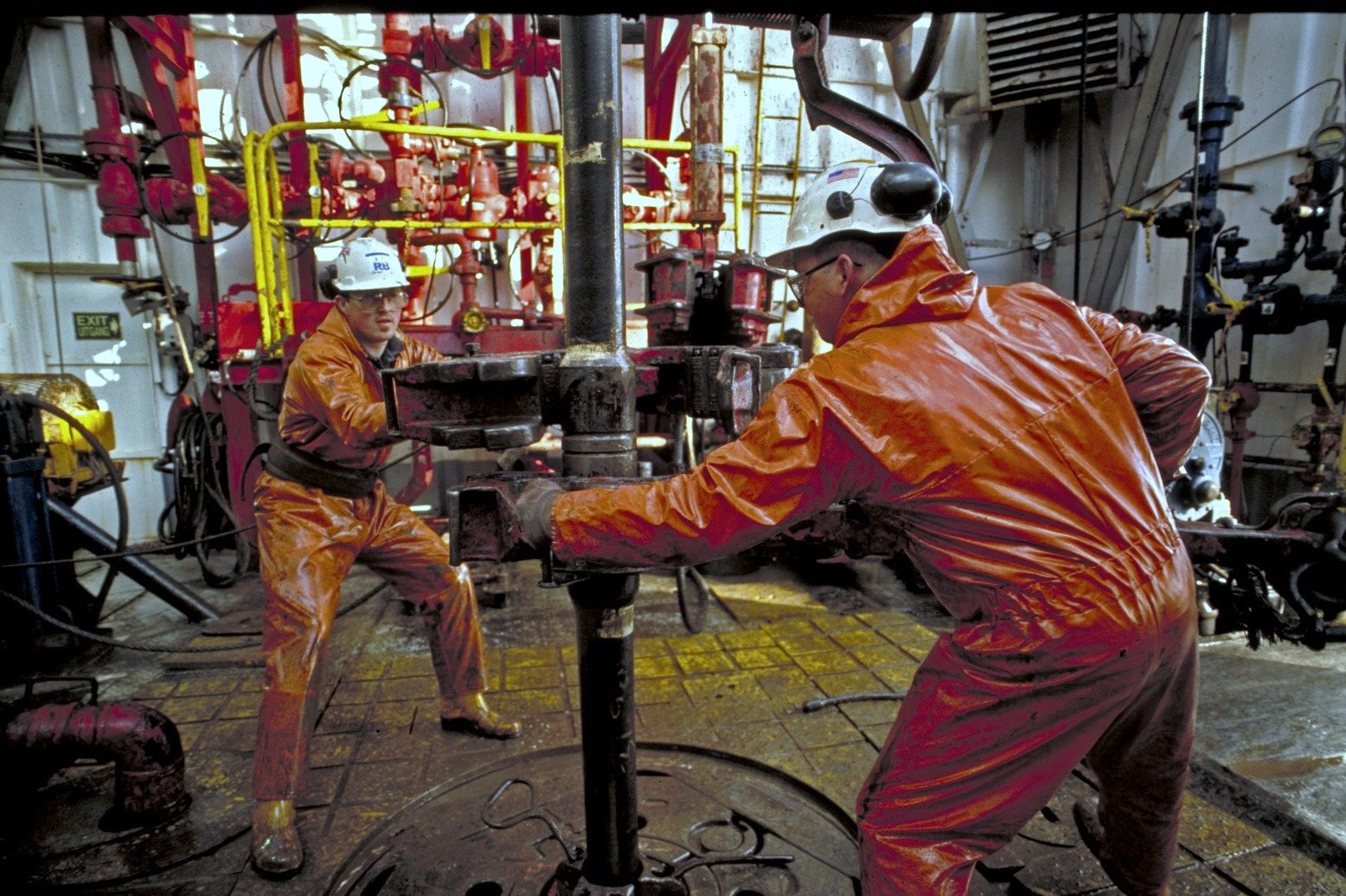 Two men in orange oilskins and helmets working at the drill pipe on the drill floor