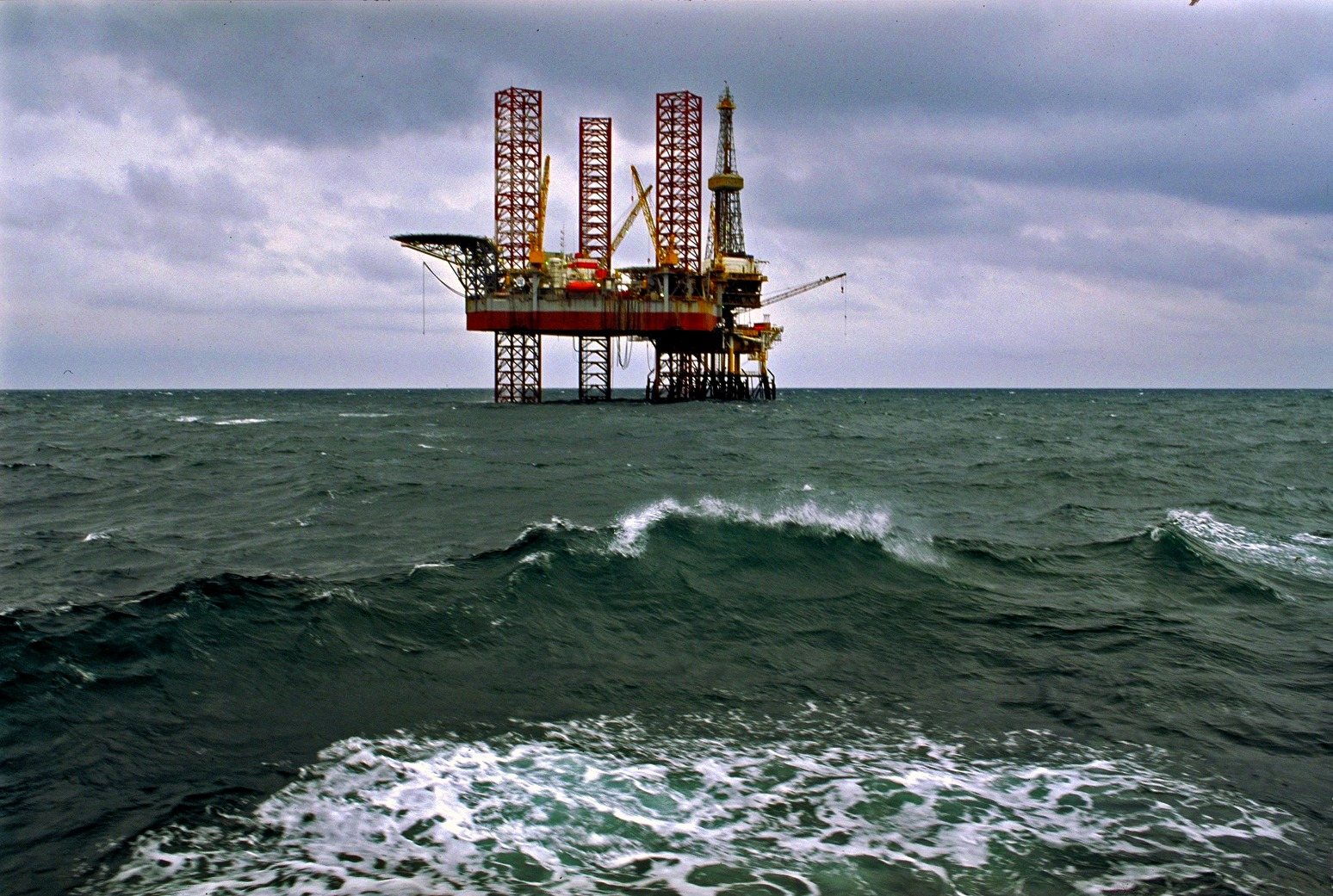 A jack-up drilling rig on the horizon of a grey, rough sea