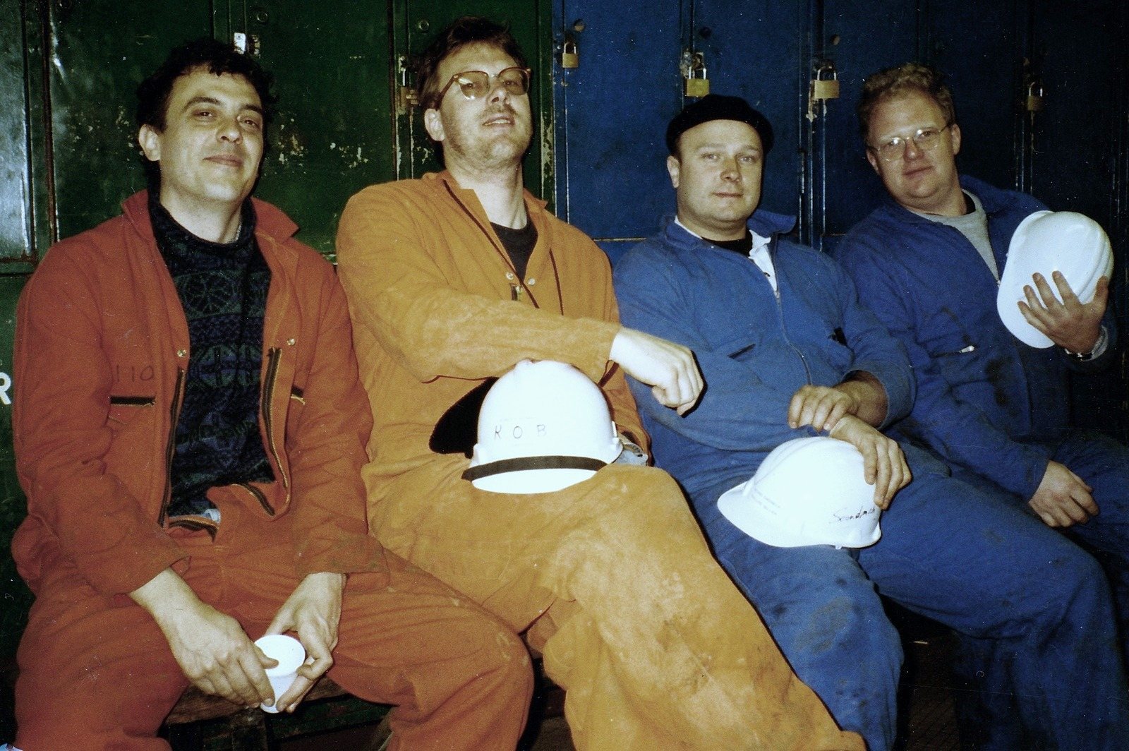 Four men in orange and blue overalls sit side by side, holding white helmets