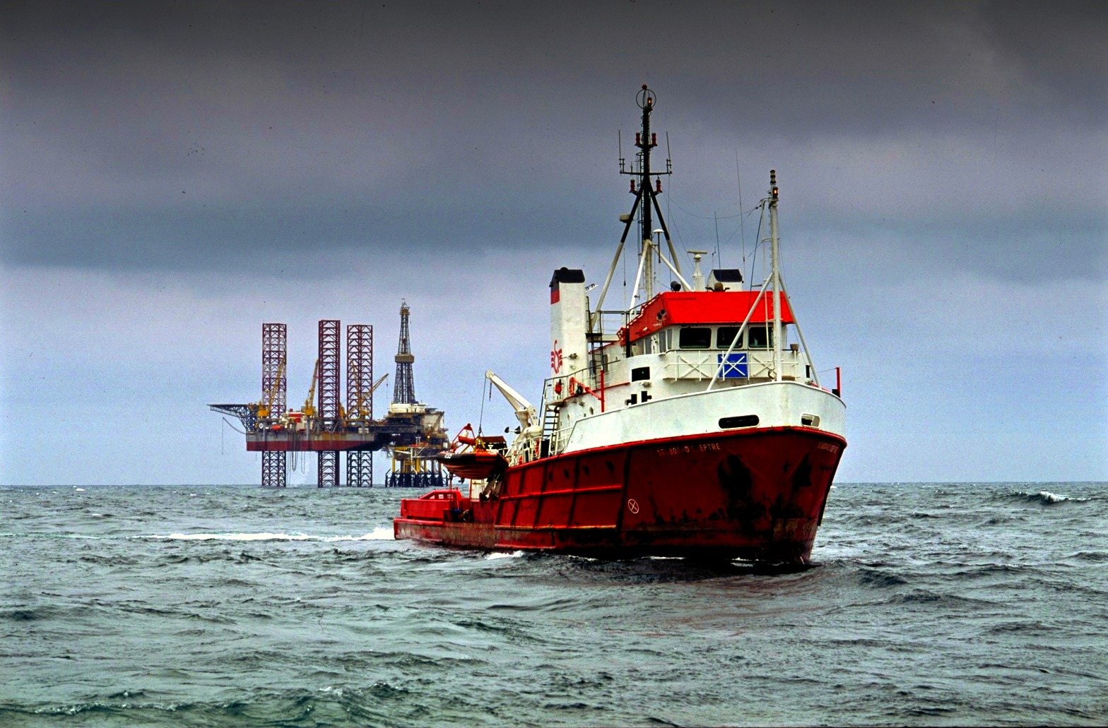 A red and white stand-by vessel in a grey sea, a drilling rig behind