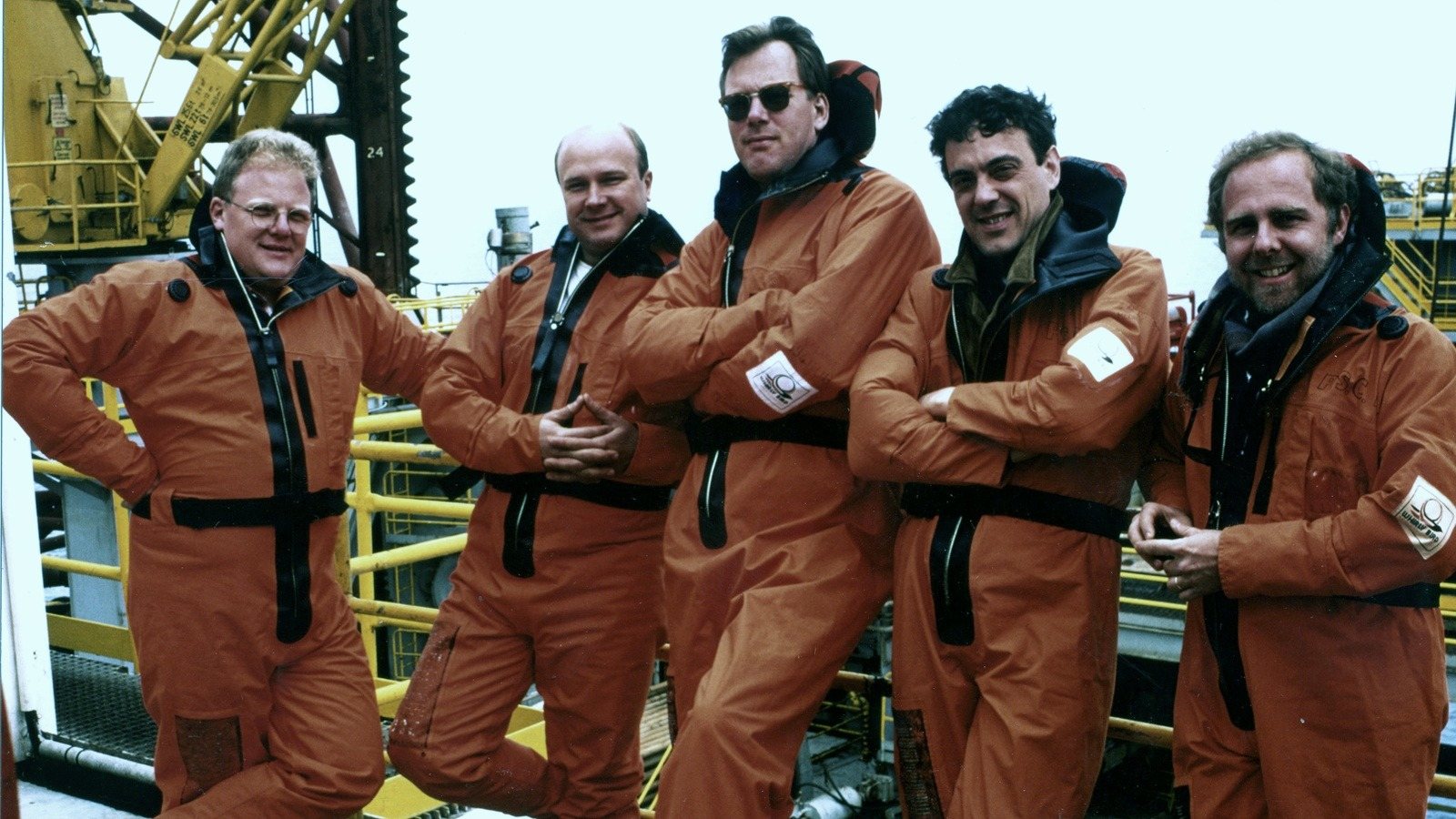 Five men in orange survival suits lean on a railing on the rig