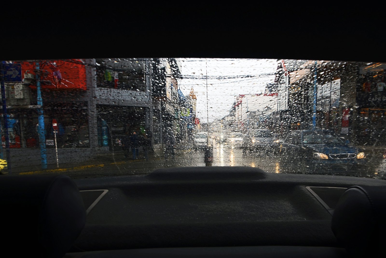 A rainy shopping street seen through the rear window of a car, raindrops on the glass, cars with headlights on