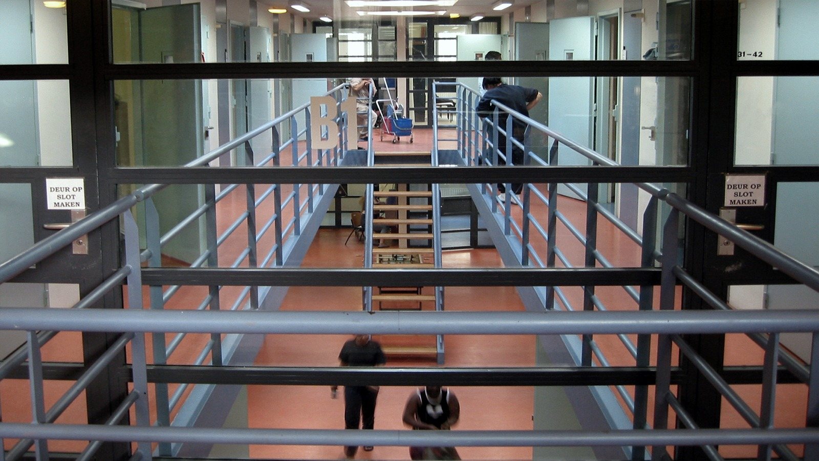 A long prison corridor seen through railings, galleries on both sides, an orange floor