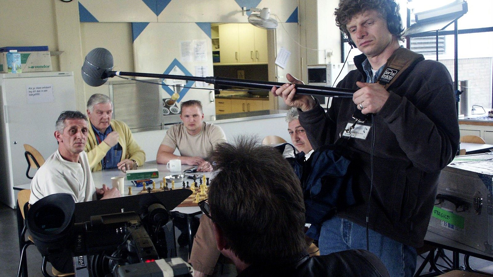 Three detainees at a table in a common room, a sound recordist with a boom and a cameraman in front