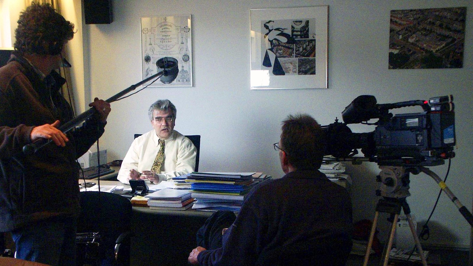 Jan van den Brand at his desk, filmed by a cameraman, a sound recordist with a boom on the left