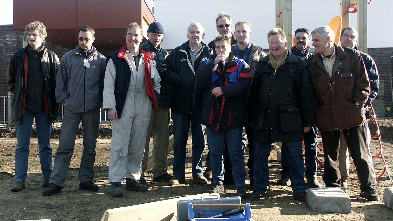 A group of about fifteen men standing on bare ground in front of a building