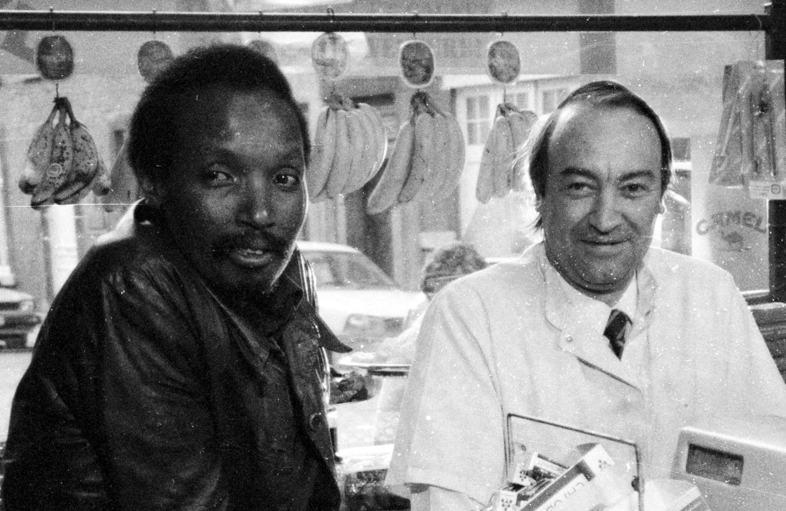 Noël N’Tunda in a leather jacket beside a smiling shopkeeper in a white coat, bananas hanging in the window behind
