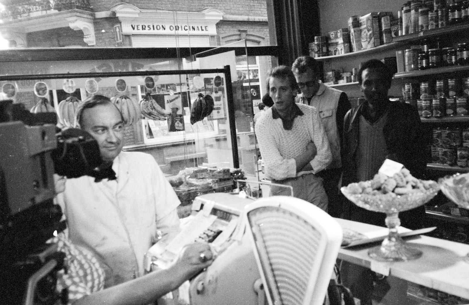 A shopkeeper at his till filmed through a shop window, a camera in the foreground and three men watching