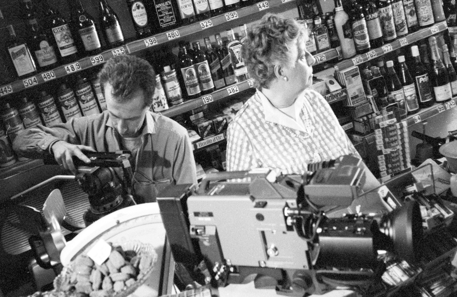 André Leplat filming with a video camera among shelves of bottles, a woman at the counter beside him