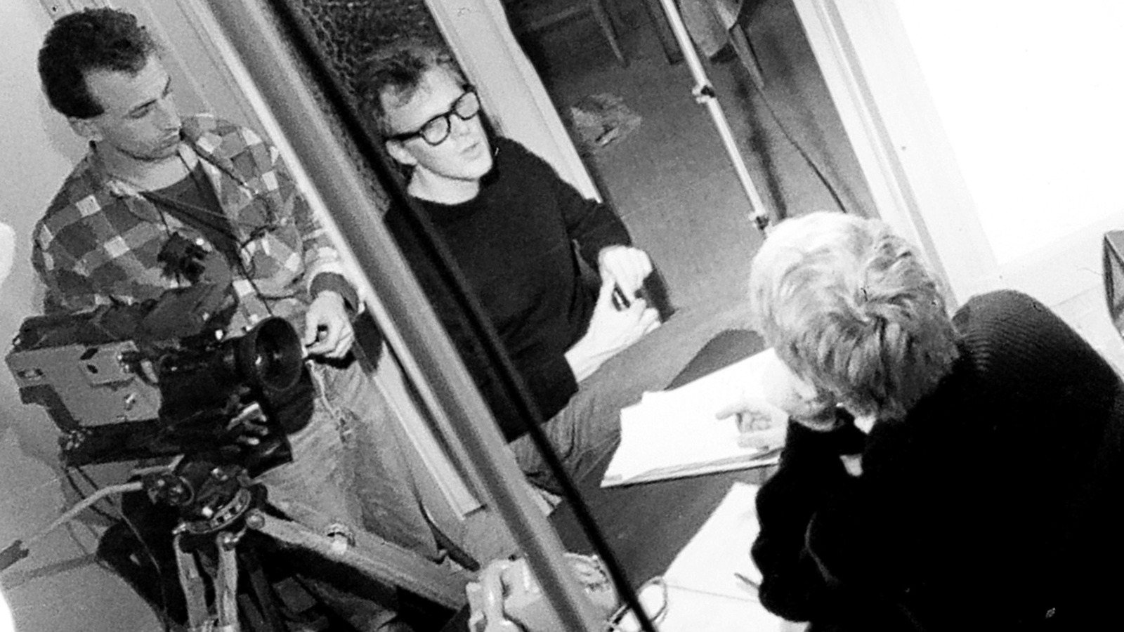 A cameraman with a video camera on a tripod, a man in glasses and a woman at a desk with papers