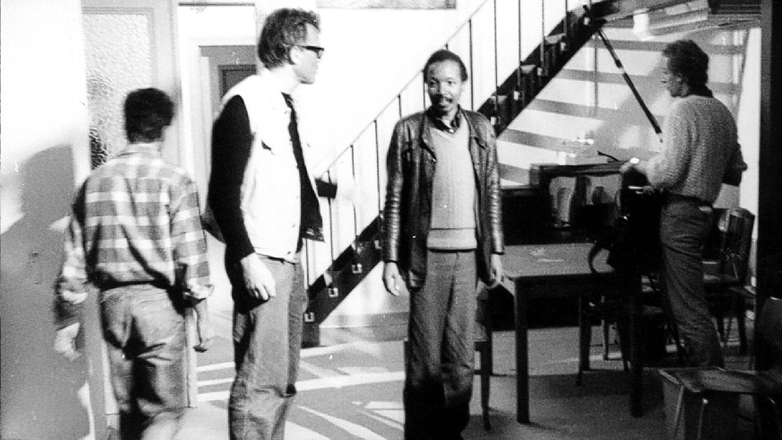 Four men in a room with a staircase, Noël N’Tunda in a leather jacket in the middle